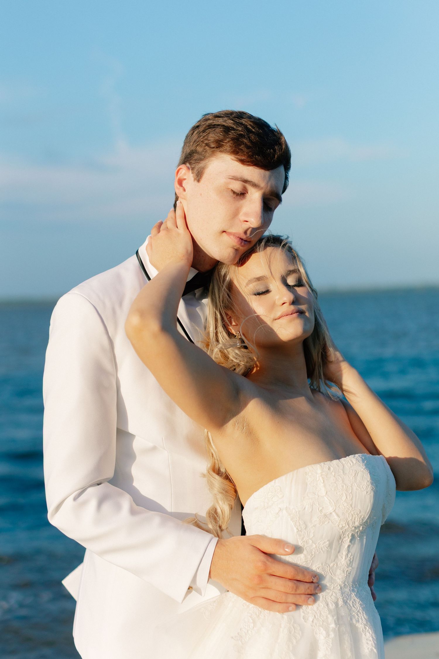 A bride and groom embrace tenderly by the ocean, both in white attire, with eyes closed in a romantic seaside wedding moment, Bella Cosa