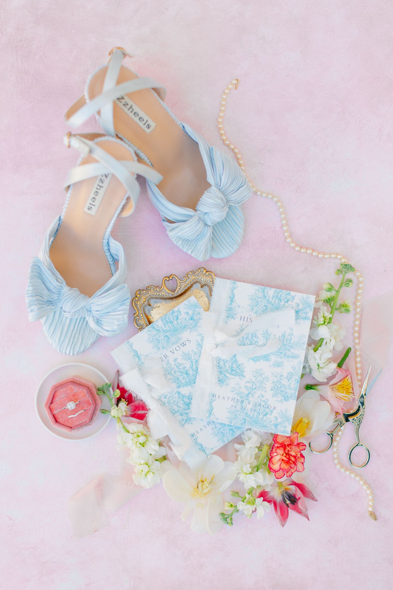 Light blue bow heels, pearl necklace, floral toile stationery, candle, and fresh flowers styled on a pink background, at Bella Cosa