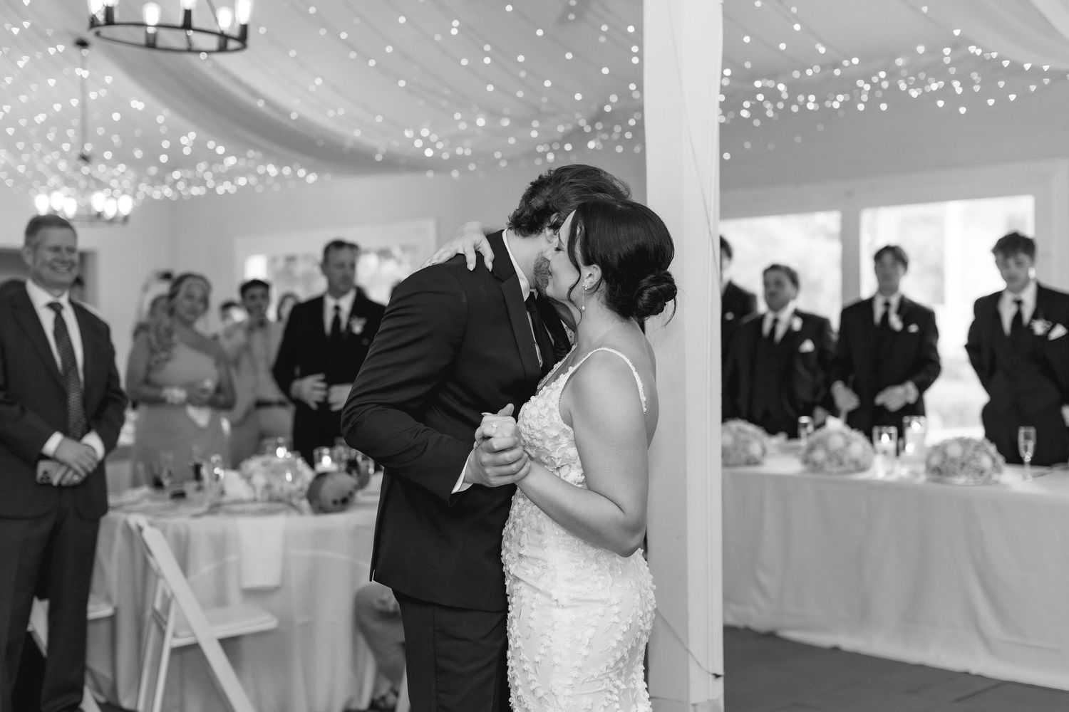 Black and white photo of bride and groom sharing first dance at elegant tented wedding reception with fairy lights.