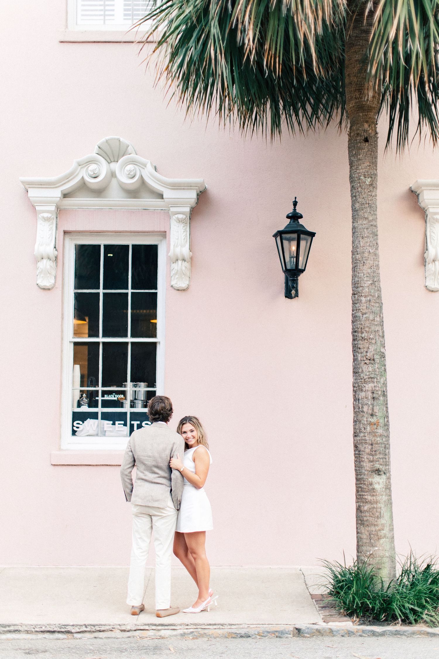 A couple stands together in front of a pink historic building with a palm tree in Charleston.