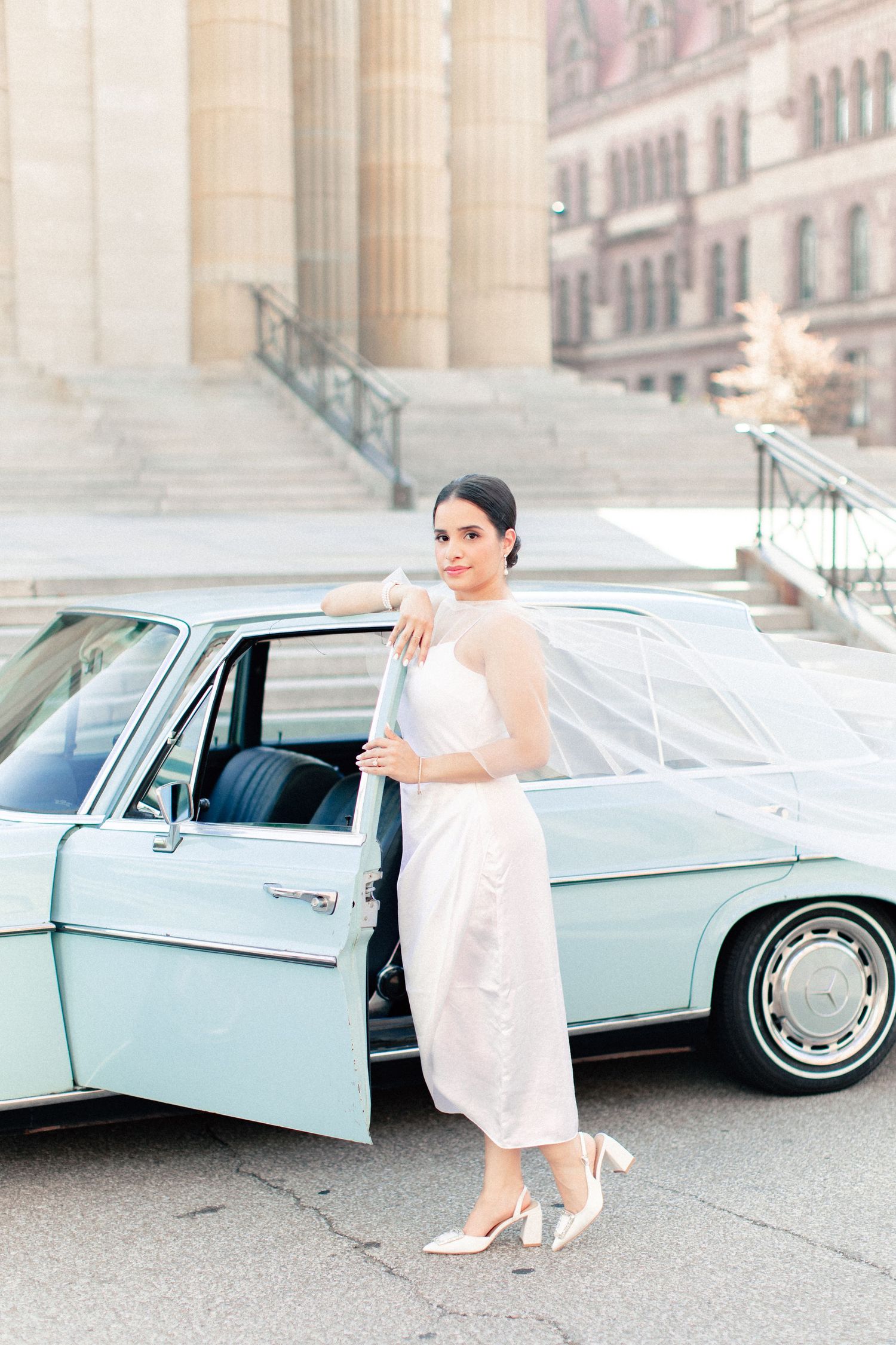 Bride in white gown poses beside a vintage mint green car in front of a classic stone building.