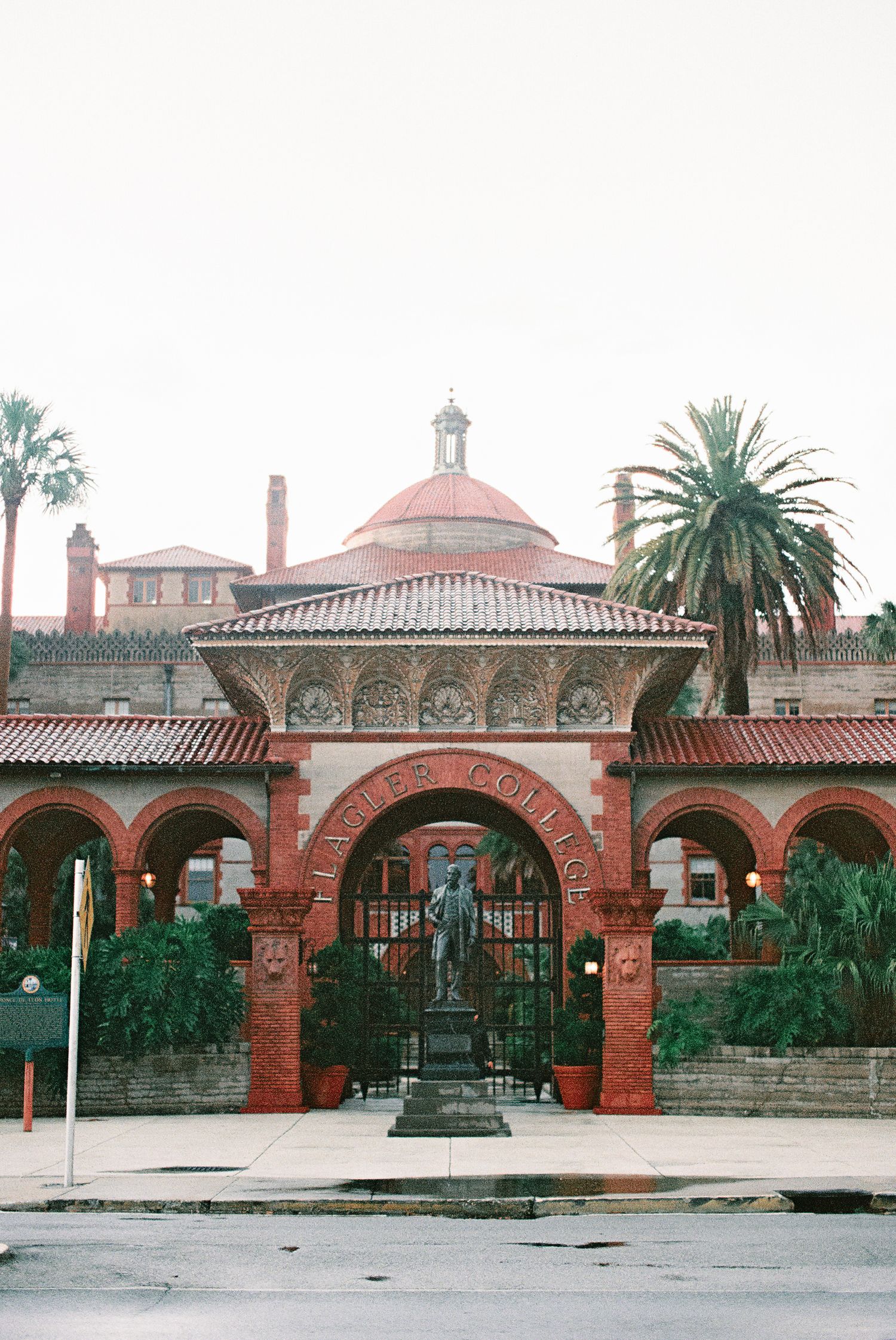Historic Flagler College entrance with ornate red brick arches, iron gates, statue, and domed building surrounded by palm trees.