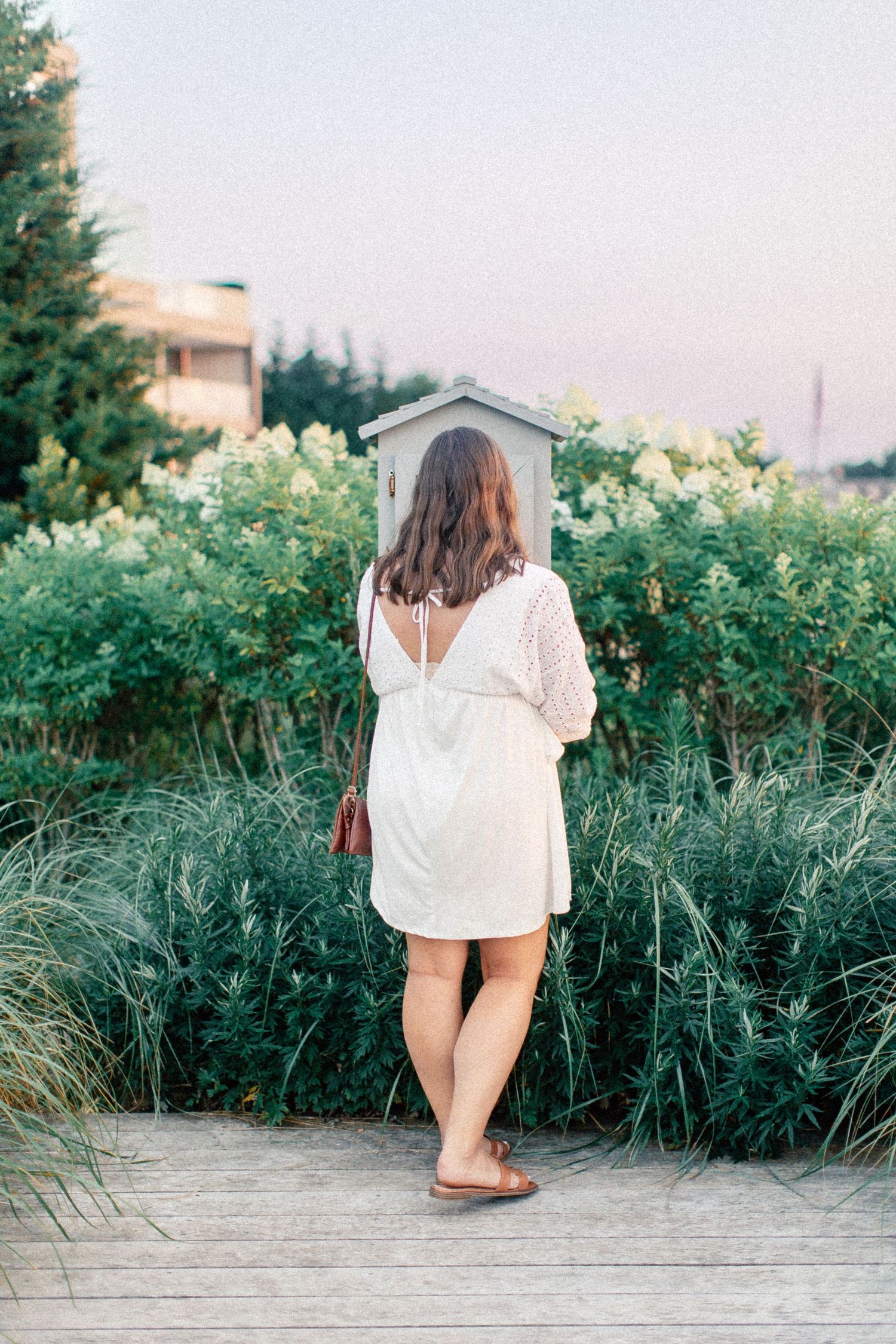 Woman in white dress standing on wooden boardwalk surrounded by lush green bushes at sunset.