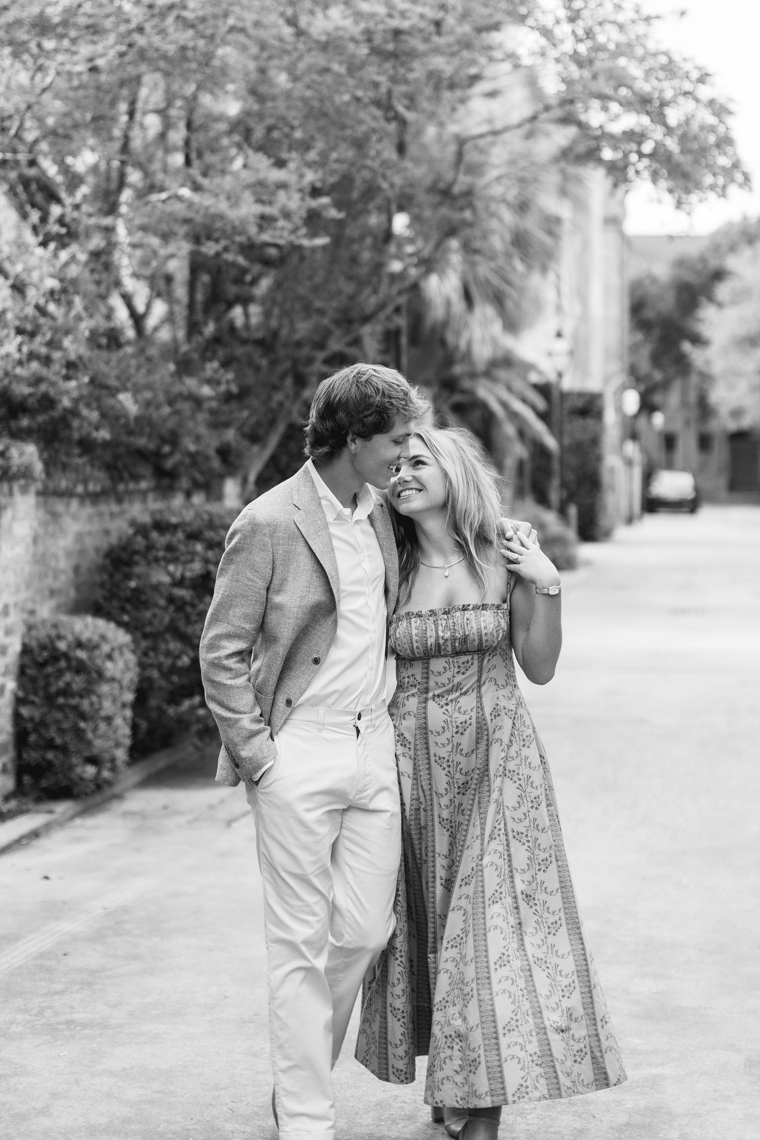 Black and white photo of a couple walking and smiling at each other on a tree-lined street.