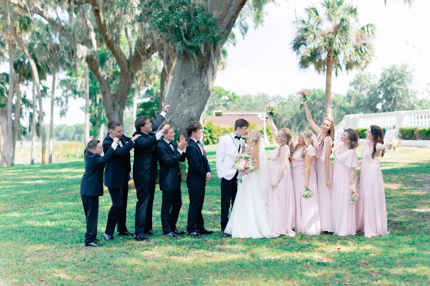 Wedding party poses outdoors under Spanish moss trees, groomsmen in black tuxedos and bridesmaids in blush pink gowns, wedding at Bella Cosa