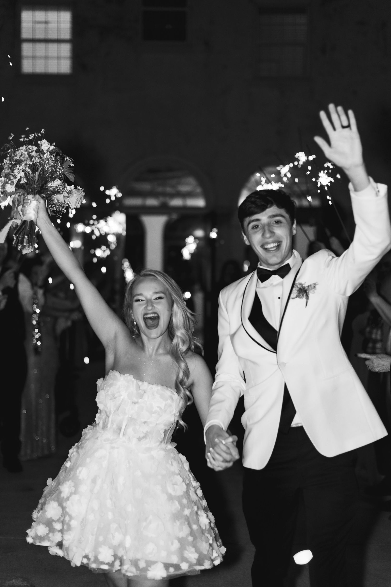 Black and white photo of joyful newlyweds celebrating their wedding exit with sparklers, bride in short floral dress holding bouquet, Florid