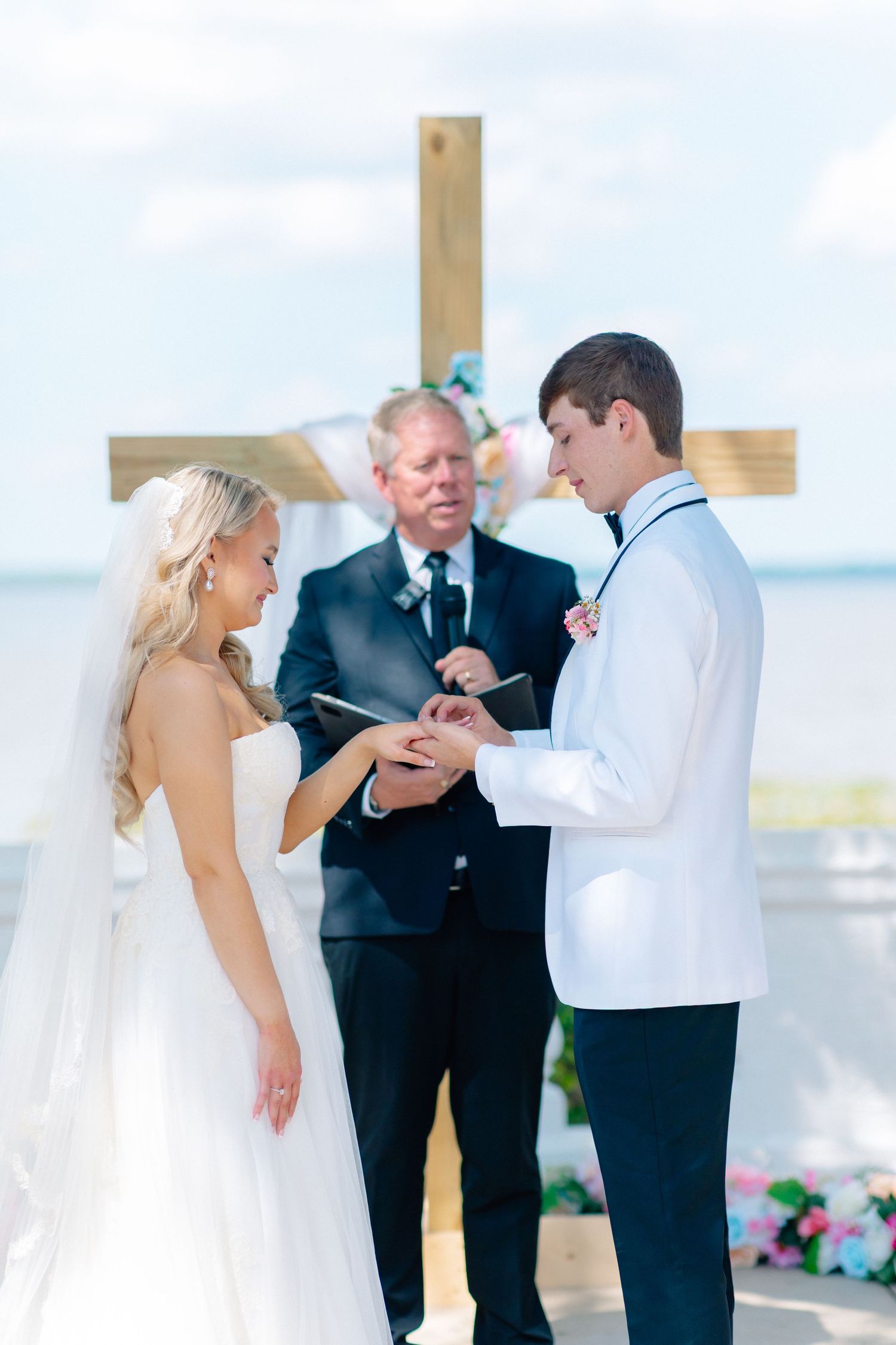 Groom placing wedding ring on bride's finger at outdoor waterfront ceremony with officiant and wooden cross backdrop, wedding at Bella Cosa