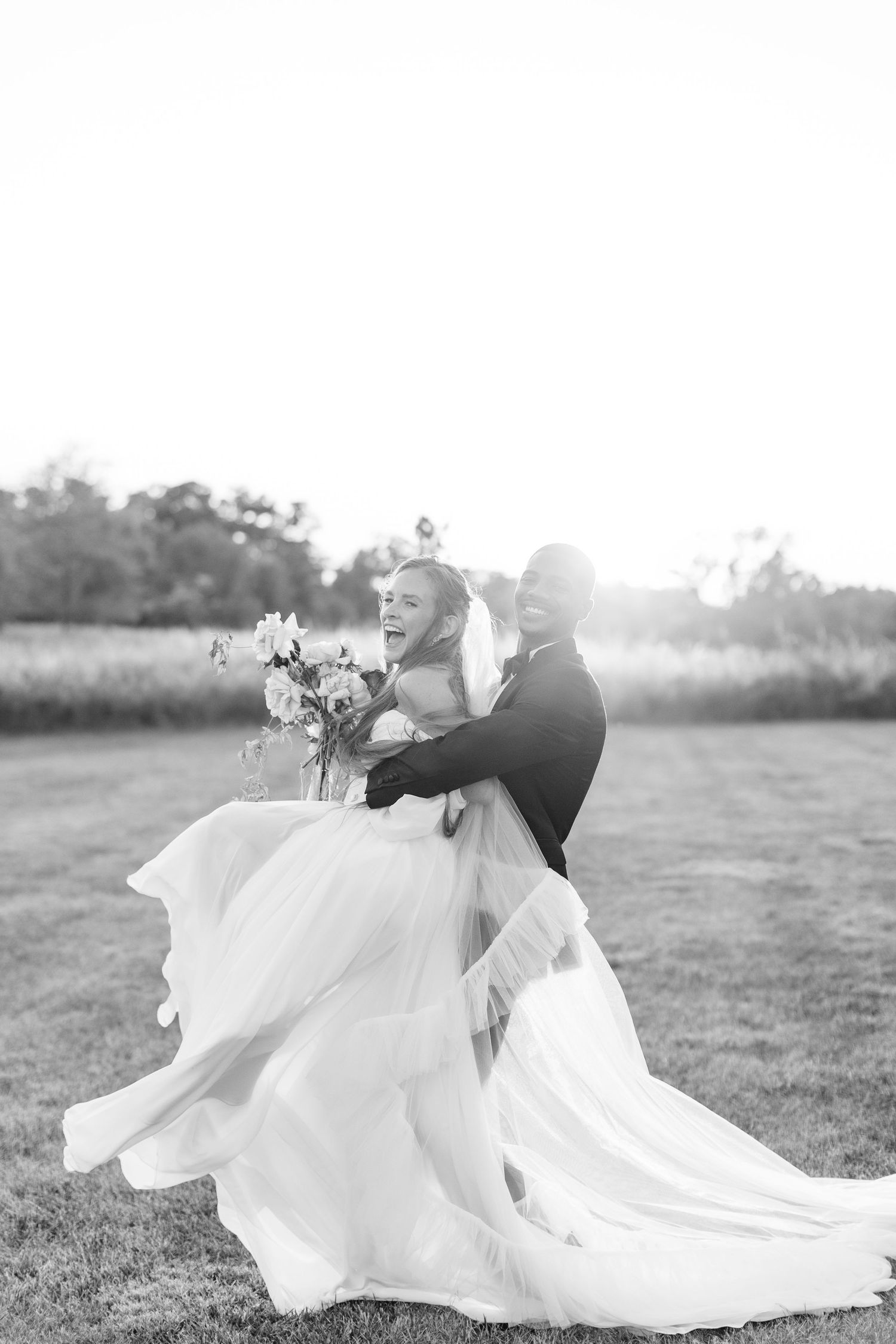 Black and white photo of groom lifting laughing bride in flowing gown in sunlit outdoor field.
