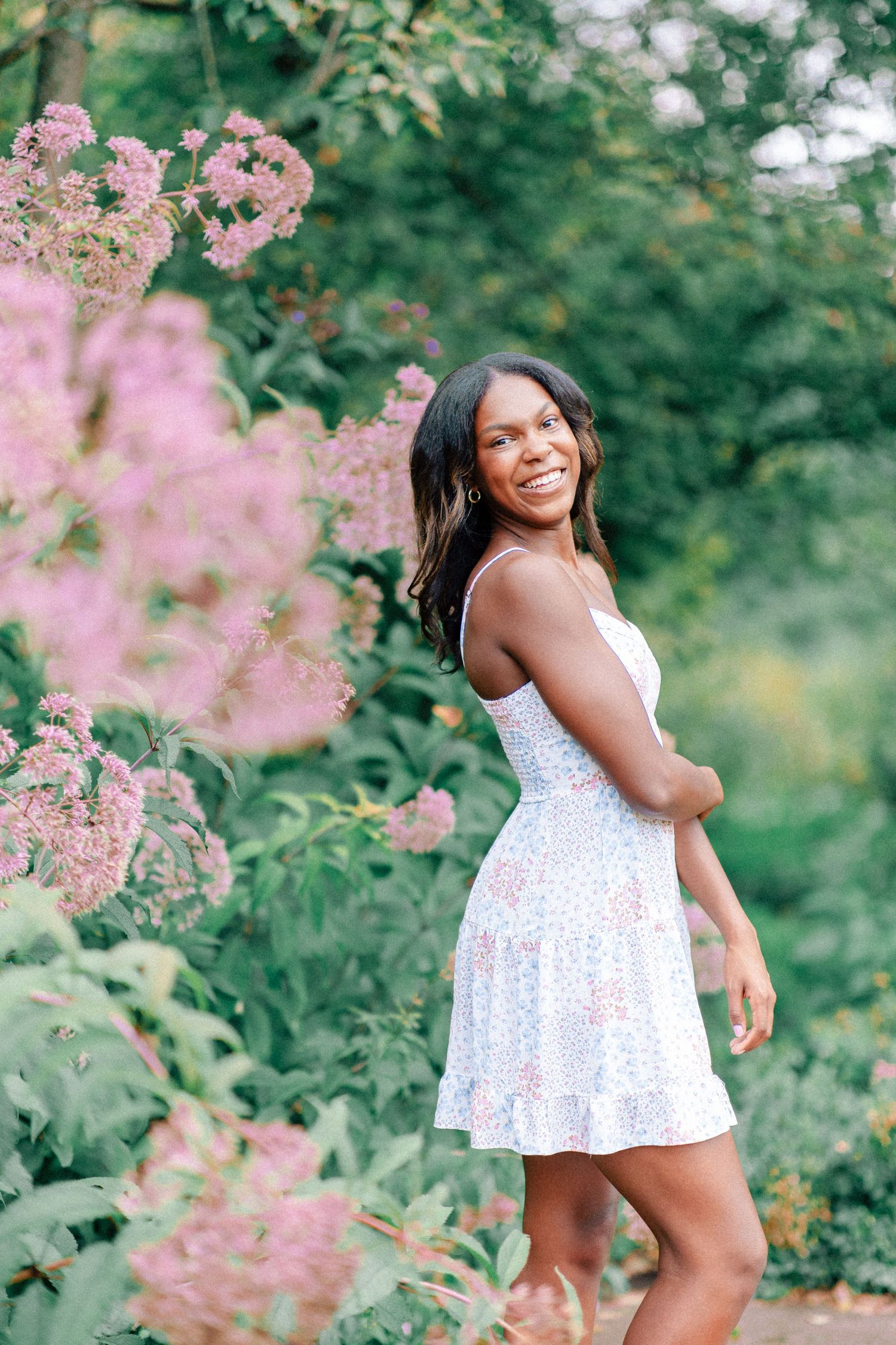 A smiling young woman in a white floral dress poses among lush pink blooms and green foliage outdoors at Ault Park