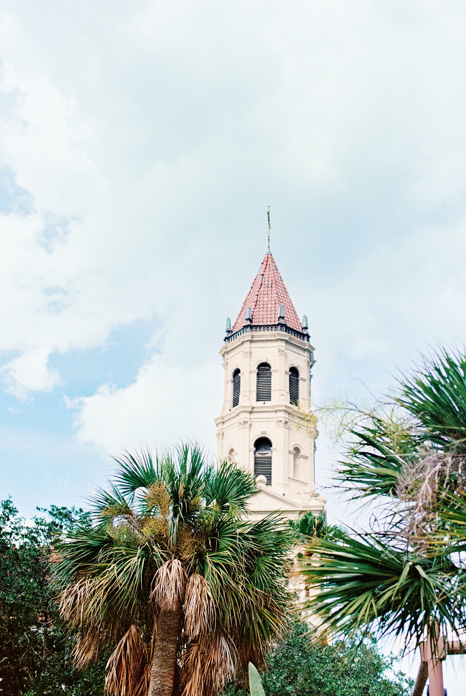 Historic church bell tower with red spire rises above lush palm trees against a cloudy blue sky.