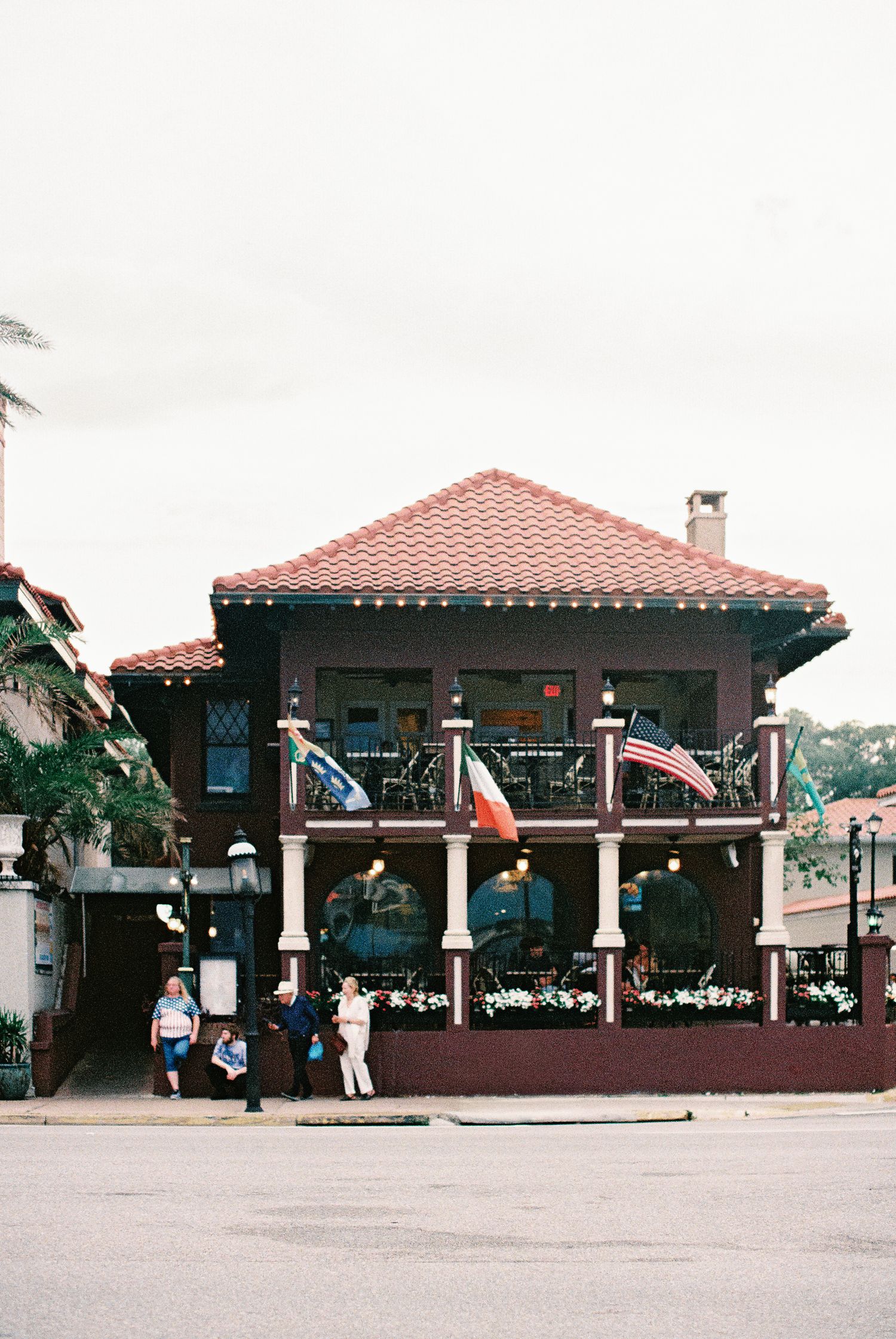 Two-story Spanish colonial building with red tile roof, flags on balcony, and pedestrians on sidewalk in historic downtown district.