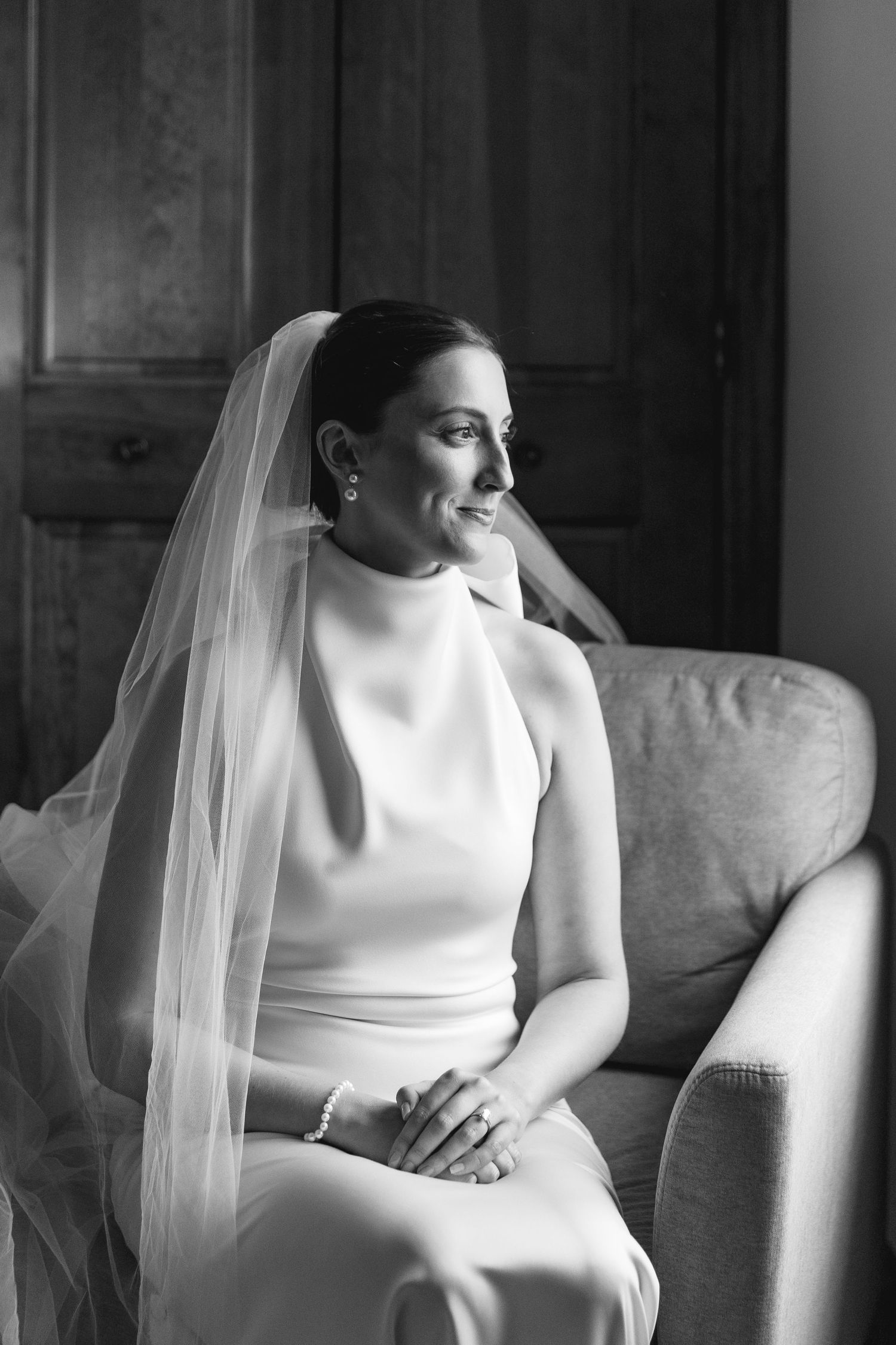 Elegant bride in sleek white gown and veil sits gracefully on sofa in black and white portrait.