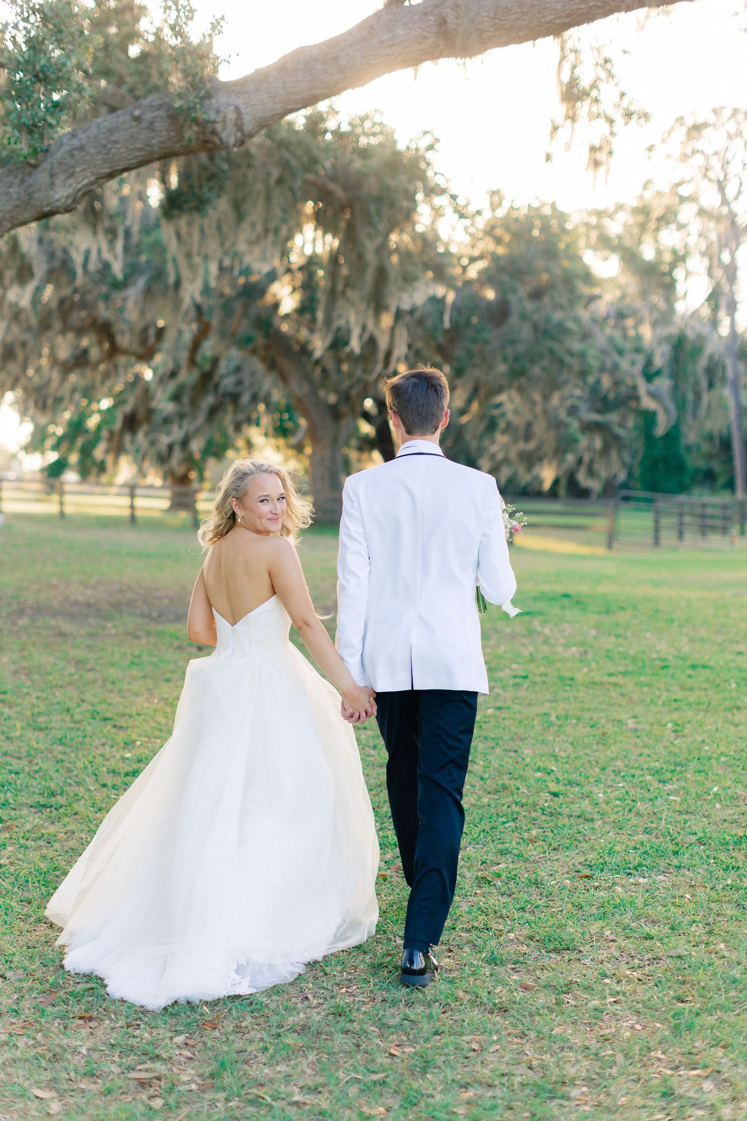 Bride in white gown looks back smiling while holding hands with groom in white jacket walking through lush green grounds, Bella Cosa
