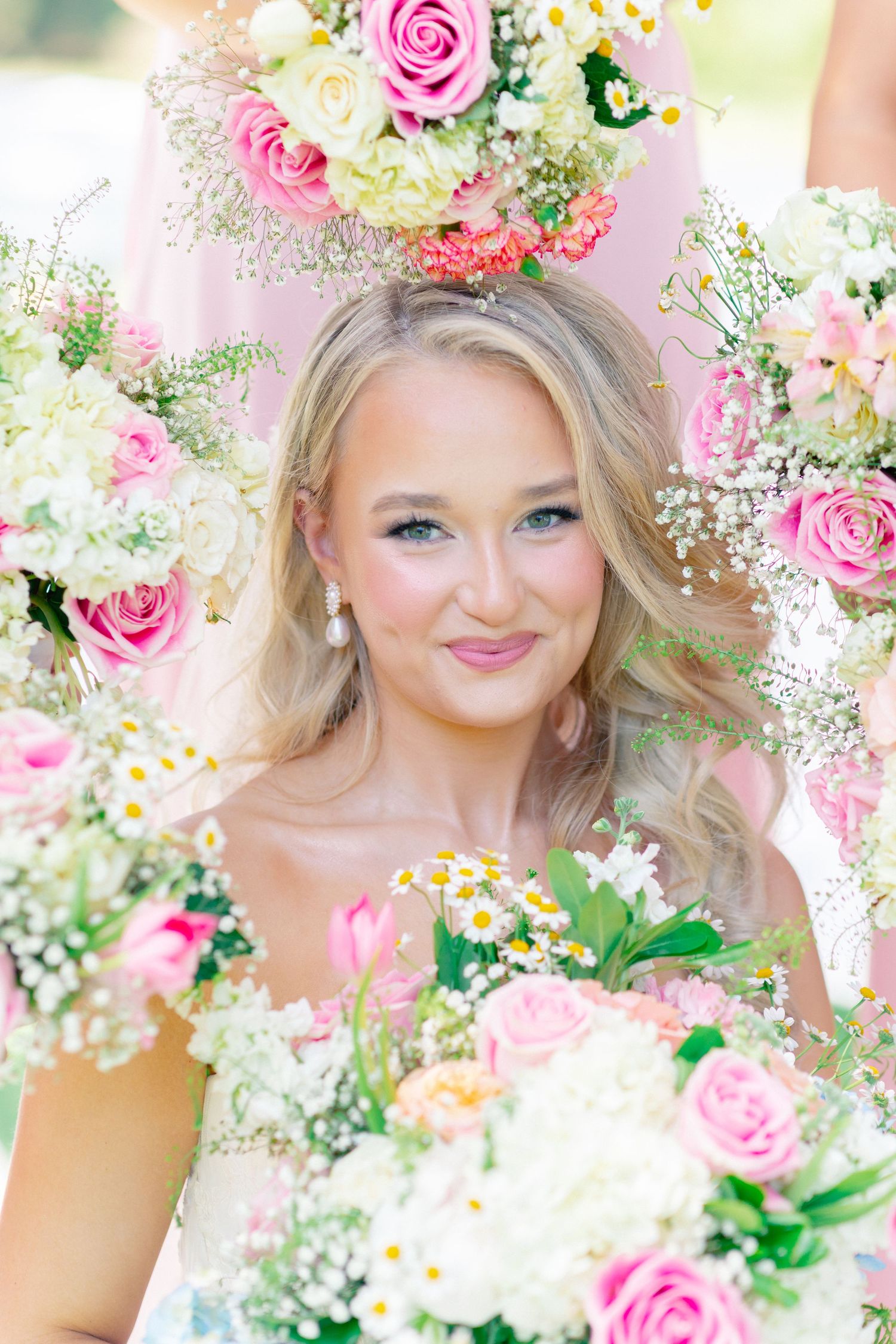 Smiling blonde bride surrounded by pink roses, white daisies, and baby's breath bouquets at an outdoor wedding ceremony, Bella Cosa