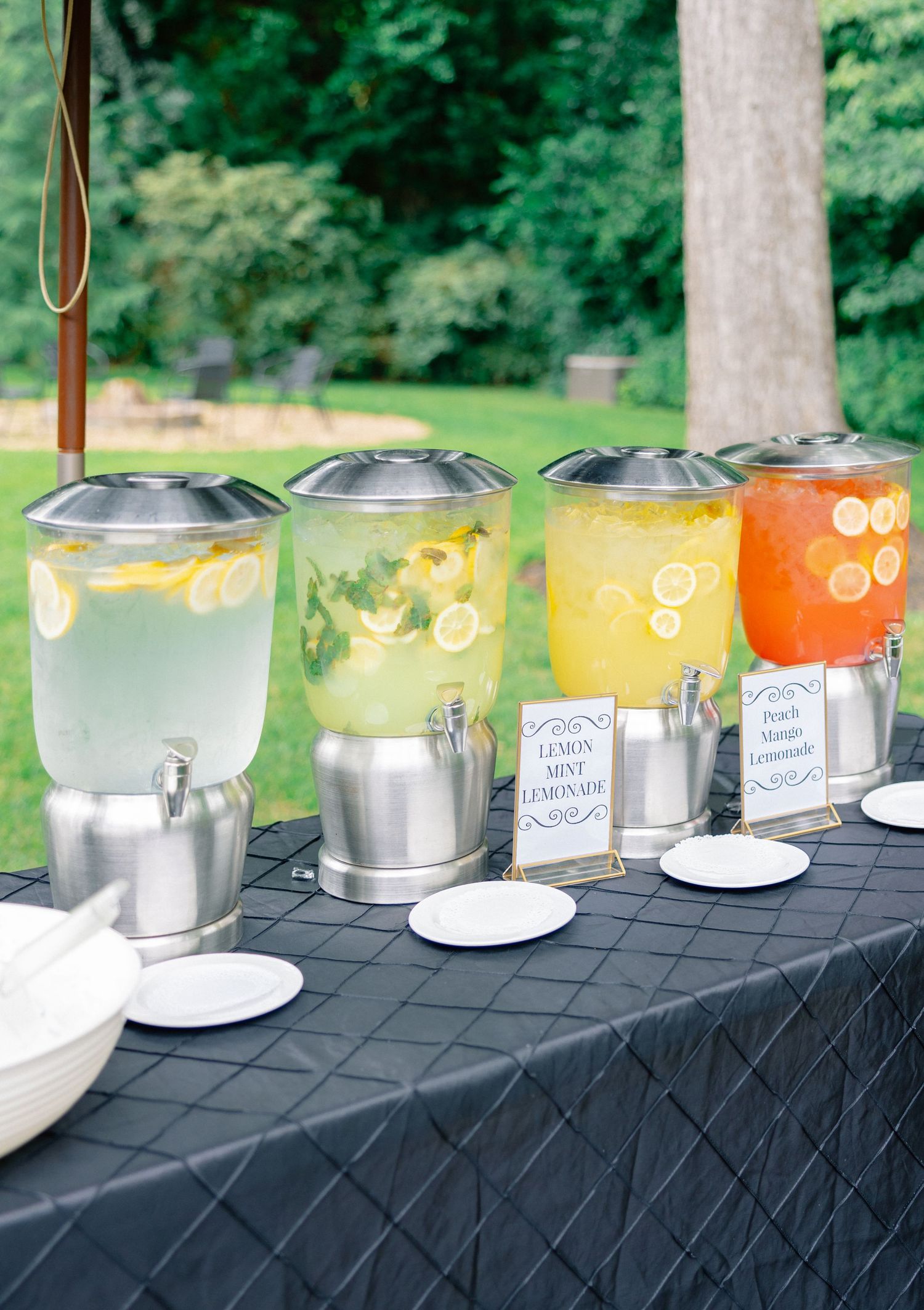 Four beverage dispensers with lemon mint lemonade and peach lemonade on a black tablecloth at an outdoor event.