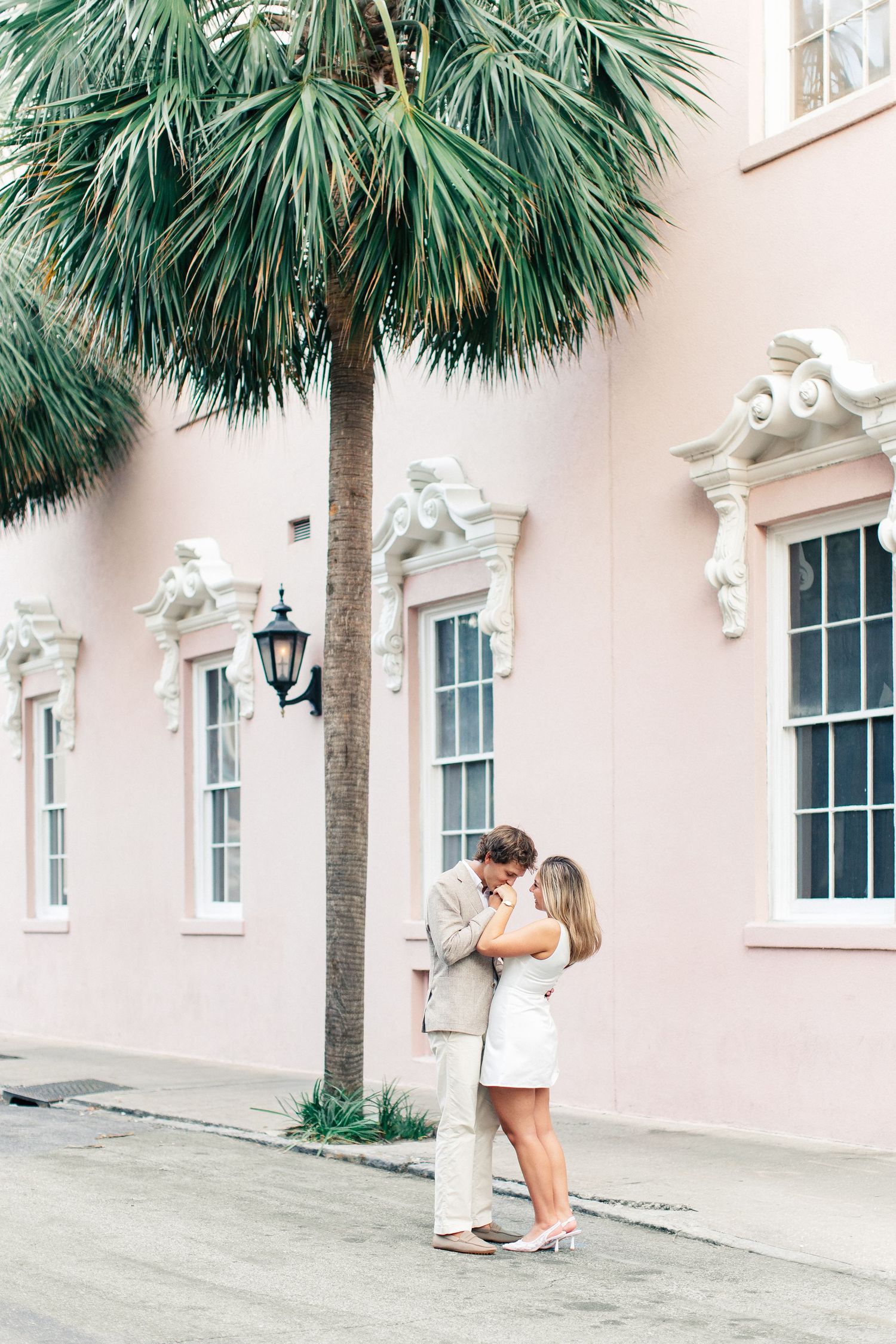 Couple sharing a romantic kiss under a palm tree beside a charming pink building in Charleston, SC.