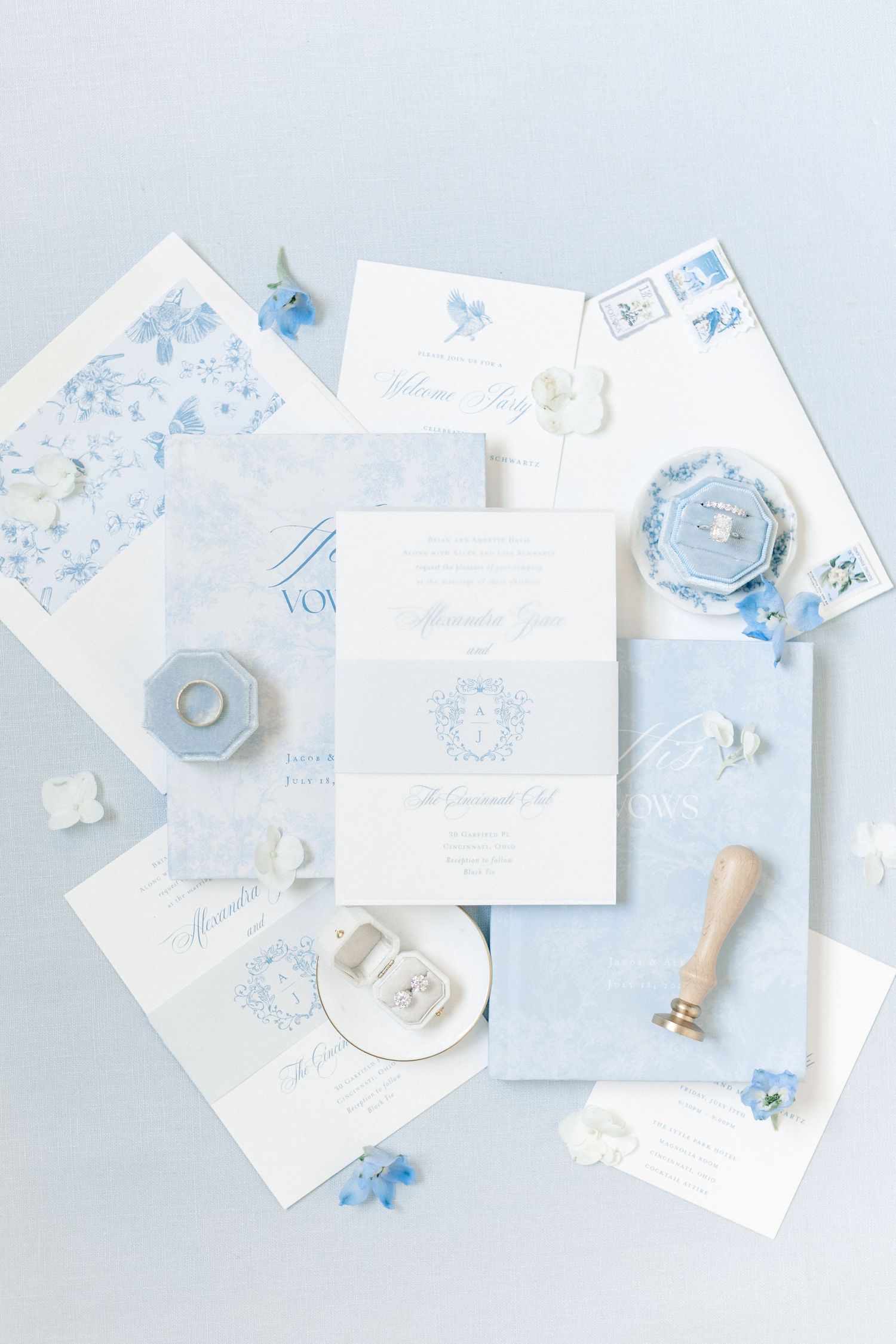 Elegant blue and white wedding stationery flat lay with floral accents, wax seal, and ring boxes on soft blue background.