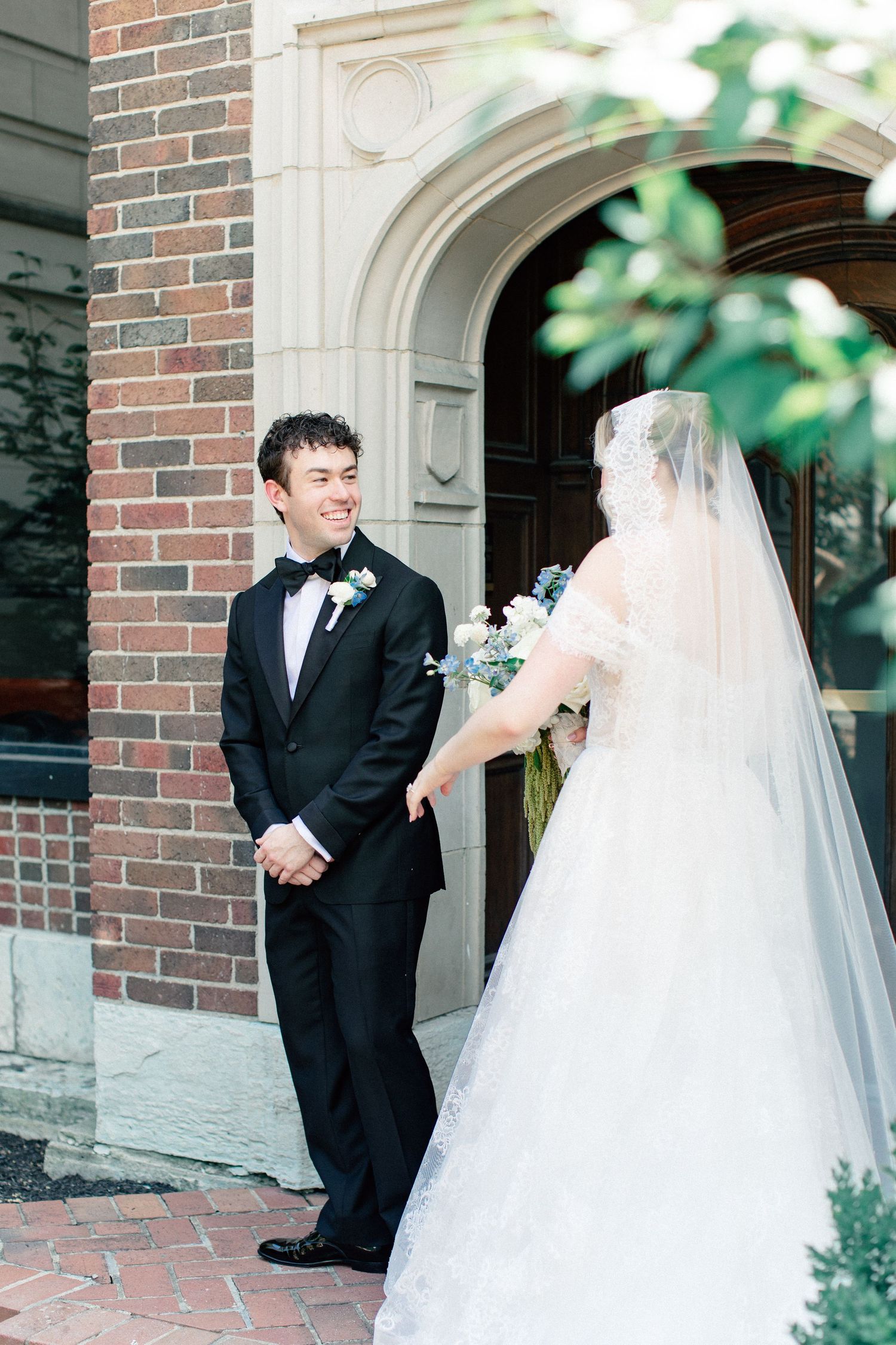 Groom in black tuxedo smiles at bride in white lace gown and veil outside brick church during first look.