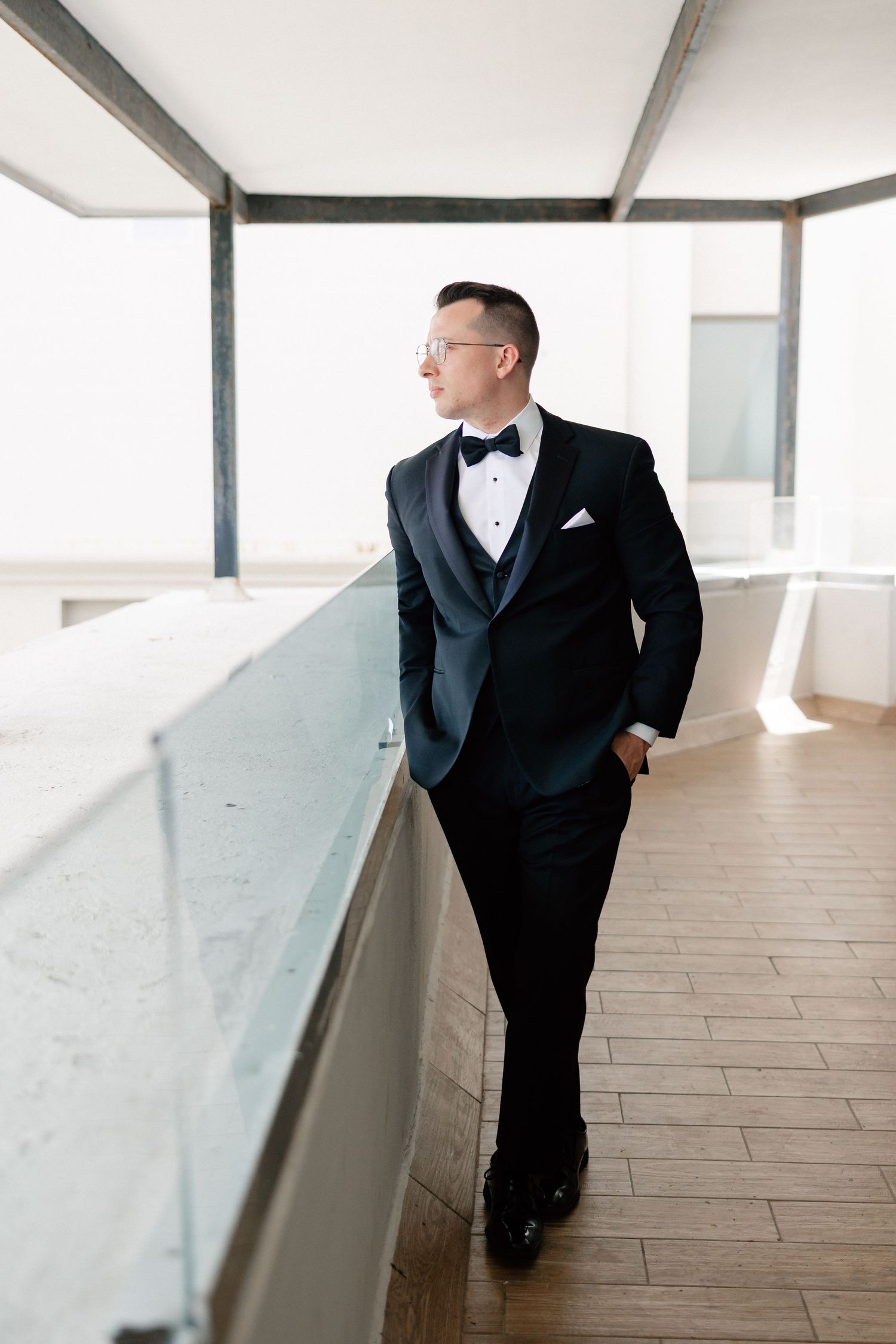 A dapper groom in a black tuxedo with bow tie leans against a railing on a sunlit balcony.