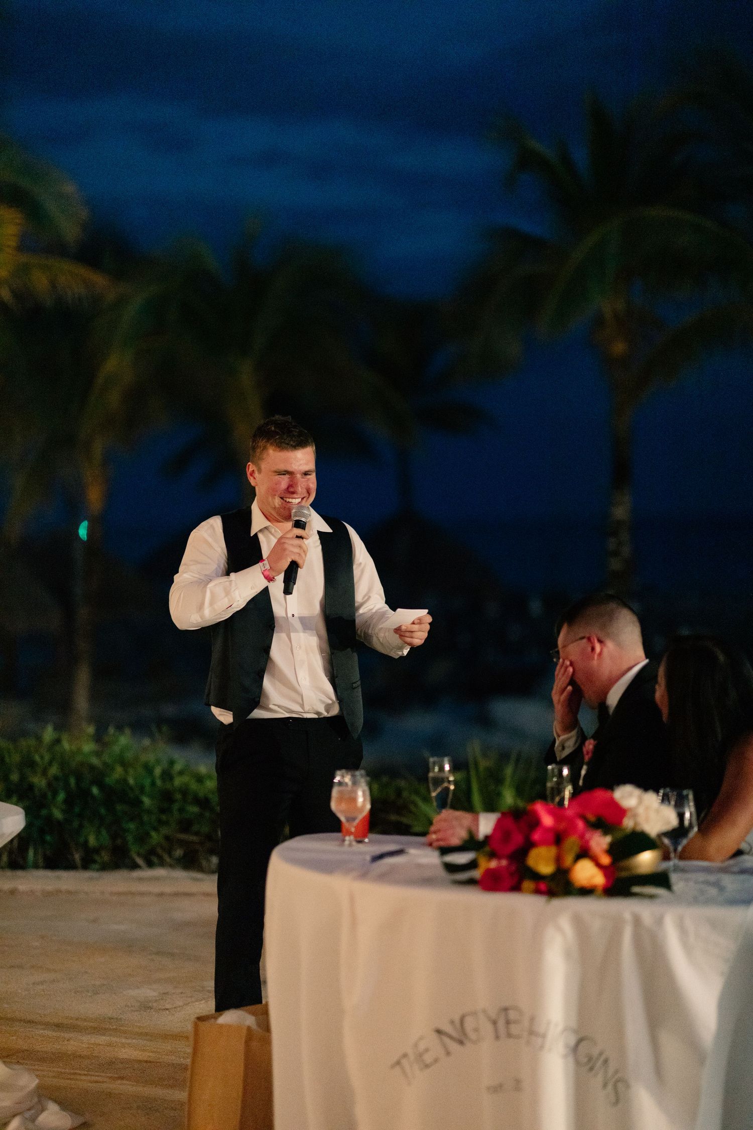 Groom giving a wedding speech outdoors at a tropical evening reception with palm trees and colorful floral centerpieces.