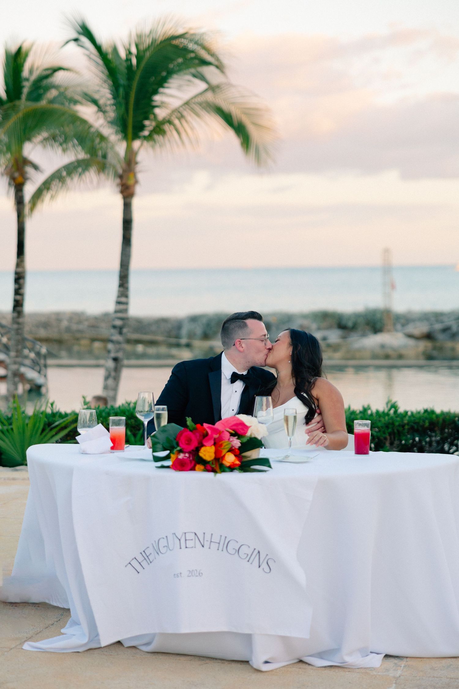 Newlyweds share a kiss at their sweetheart table with tropical palm trees and ocean backdrop at a destination wedding.