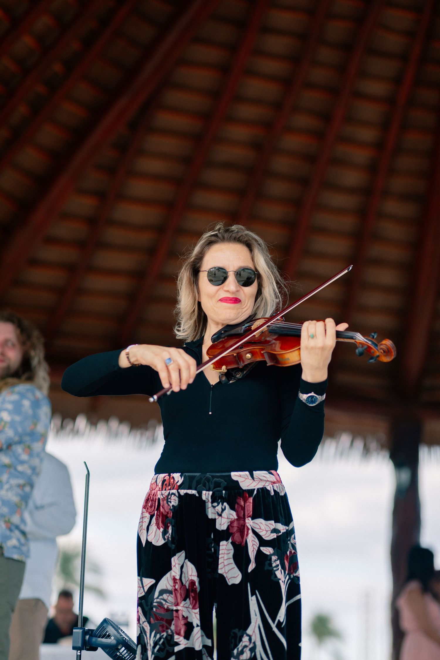 Woman playing violin outdoors under a thatched roof gazebo, wearing sunglasses and floral skirt.