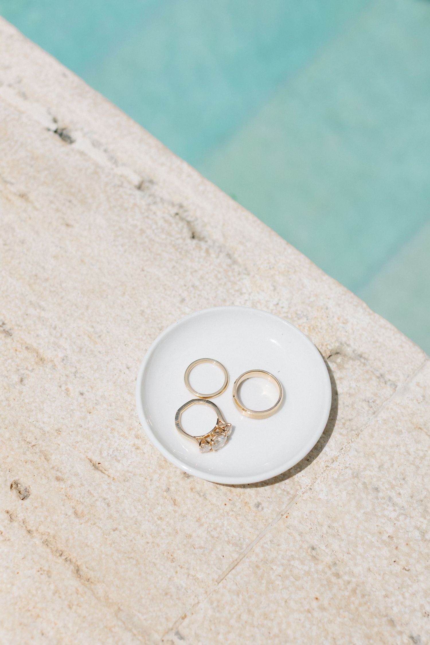 Three gold rings on a white ceramic dish by a turquoise pool edge in bright sunlight.