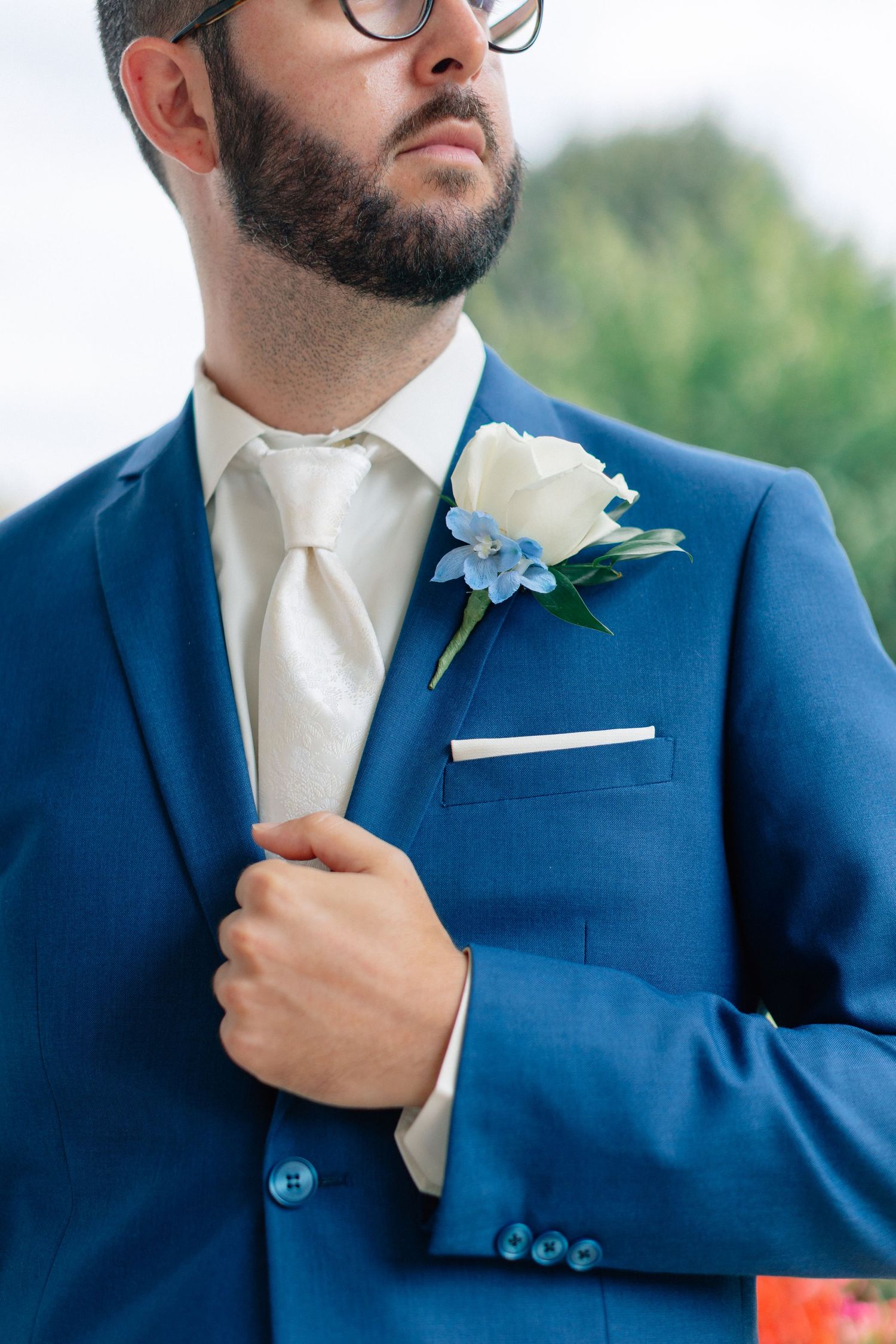 Groom in blue suit with white rose boutonniere and cream tie adjusting jacket lapel outdoors.