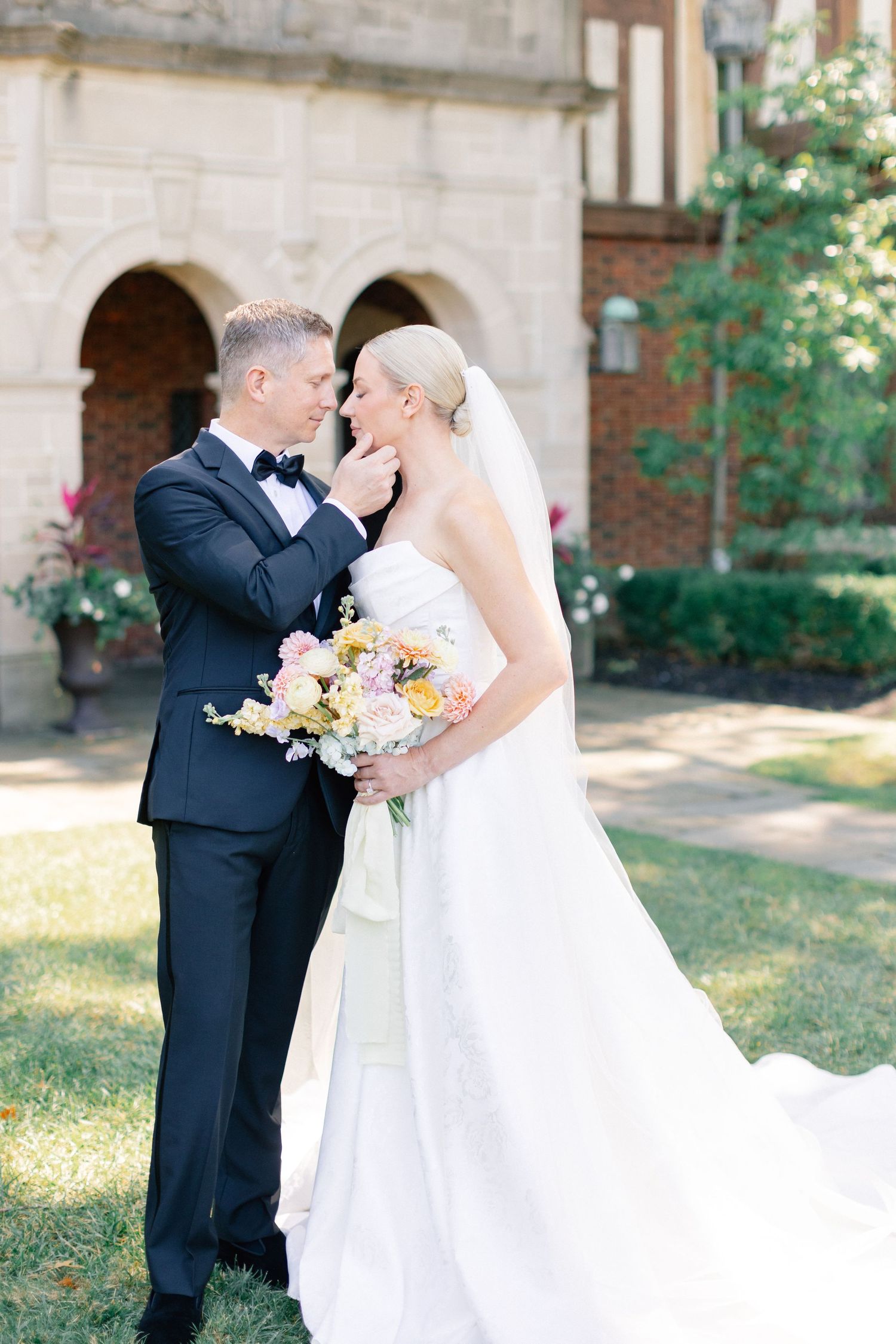 Groom tenderly cups bride's face as they stand close together outside a historic brick arched building.