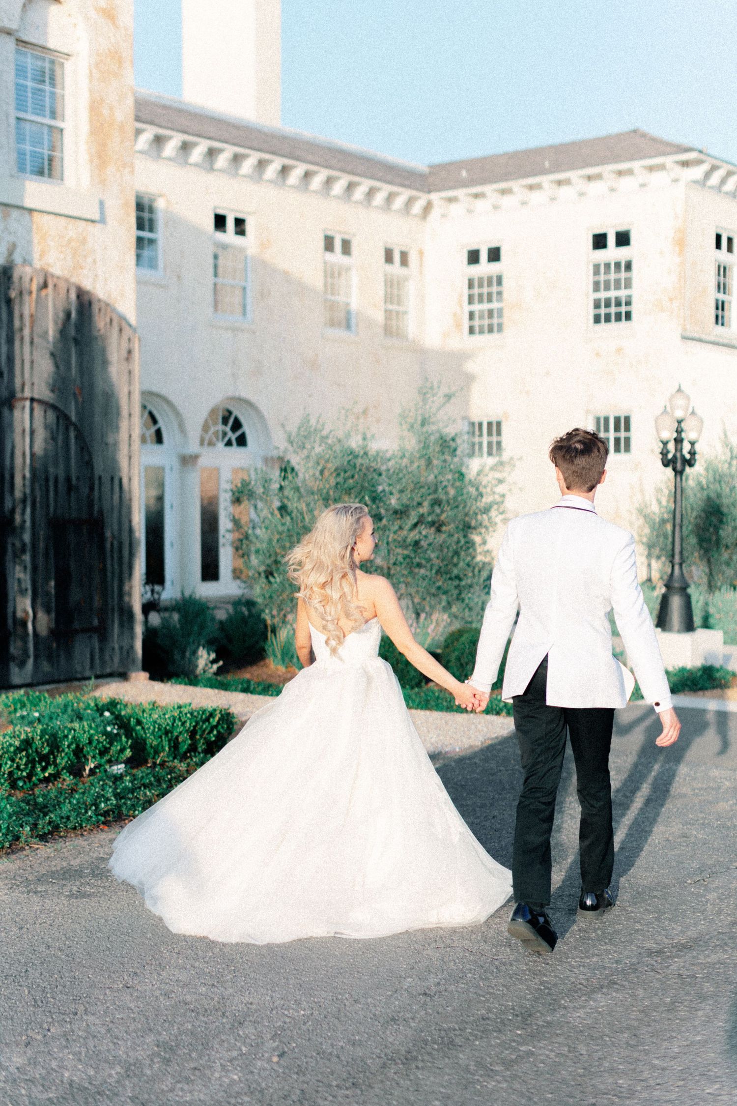 Bride in white ballgown and groom in white tuxedo hold hands walking toward elegant stone mansion.
