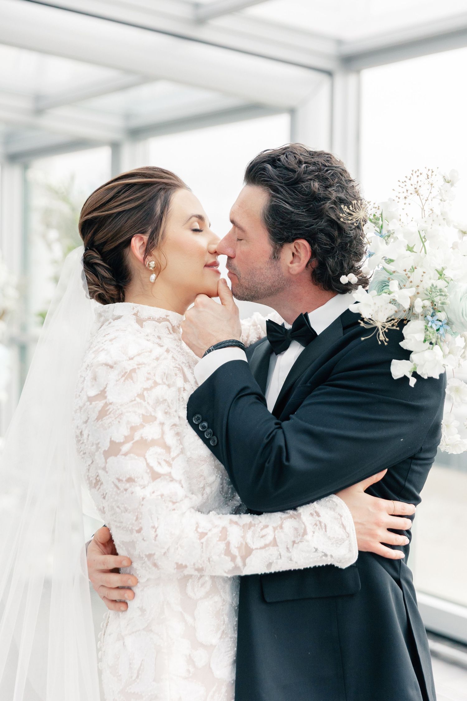 Bride and groom share a romantic kiss in elegant wedding attire, with lace dress and black tuxedo, holding white floral bouquet.