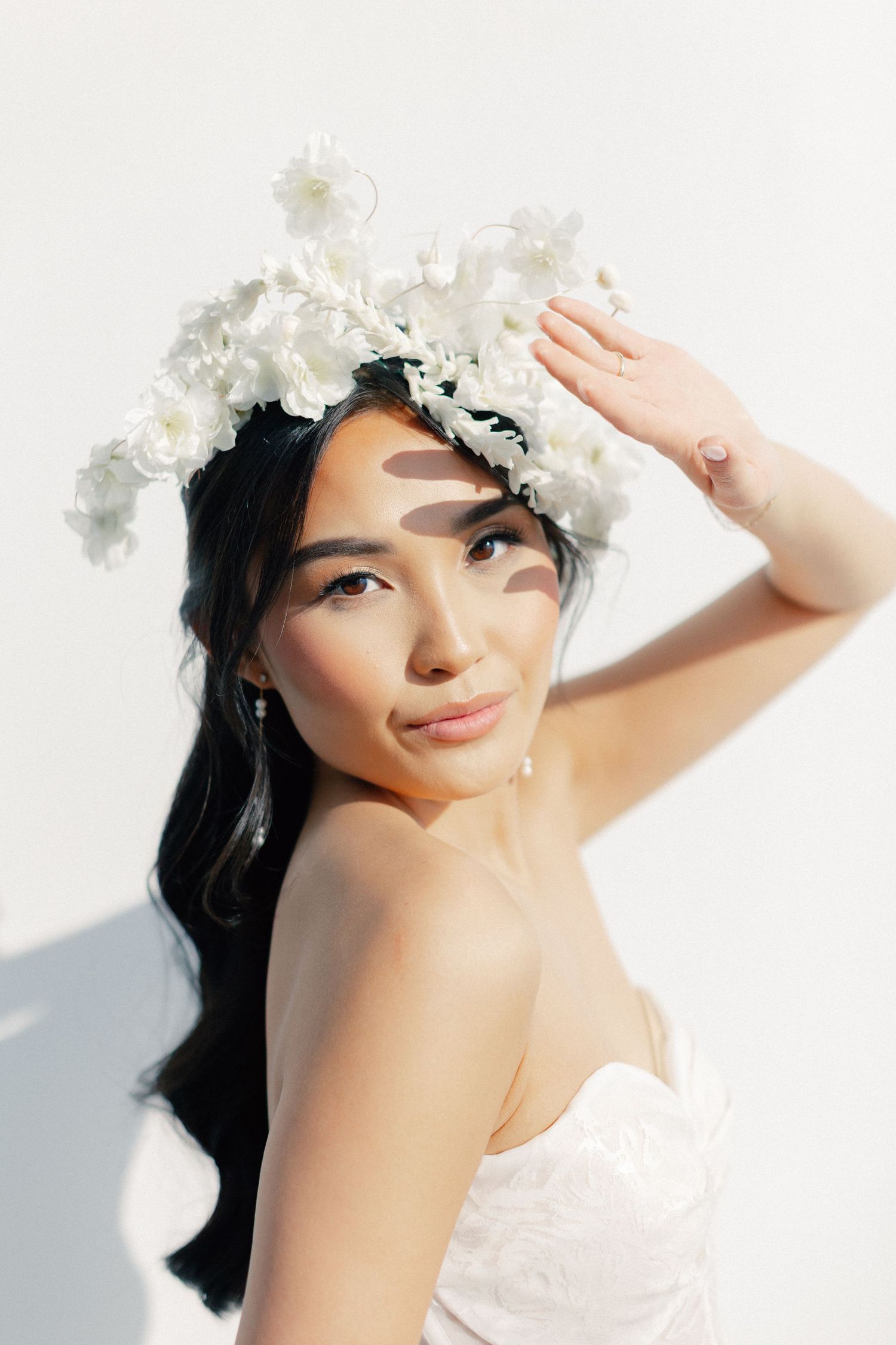 Young woman in white floral crown and strapless dress poses gracefully against a bright white background.