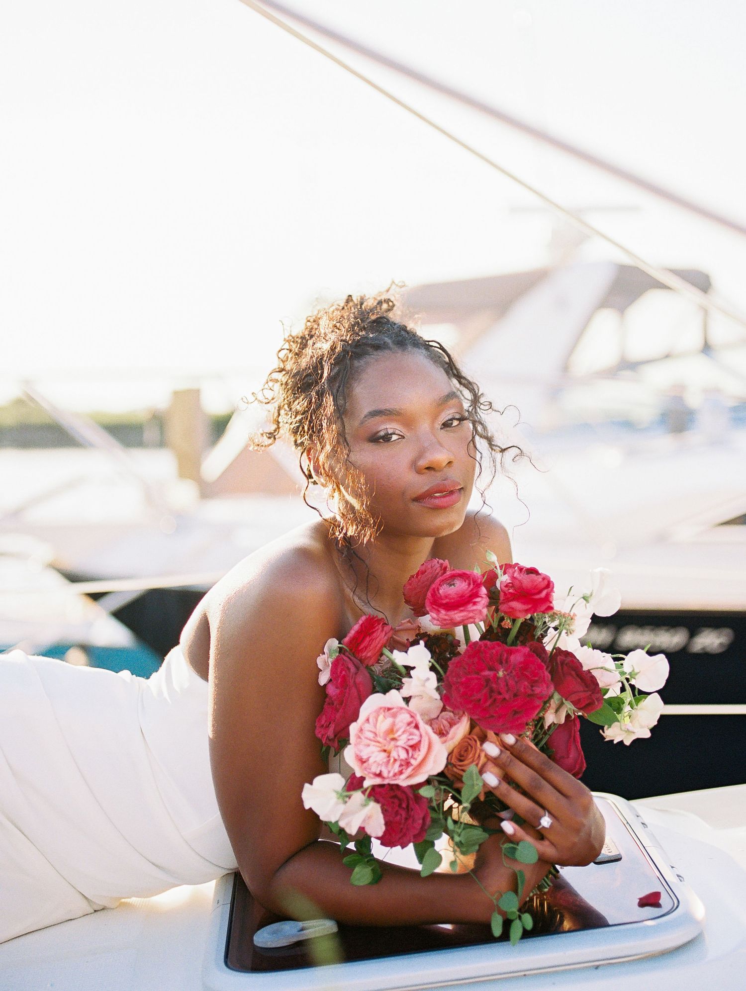 Bride holding red and pink floral bouquet on a yacht at golden hour, wearing a strapless gown.