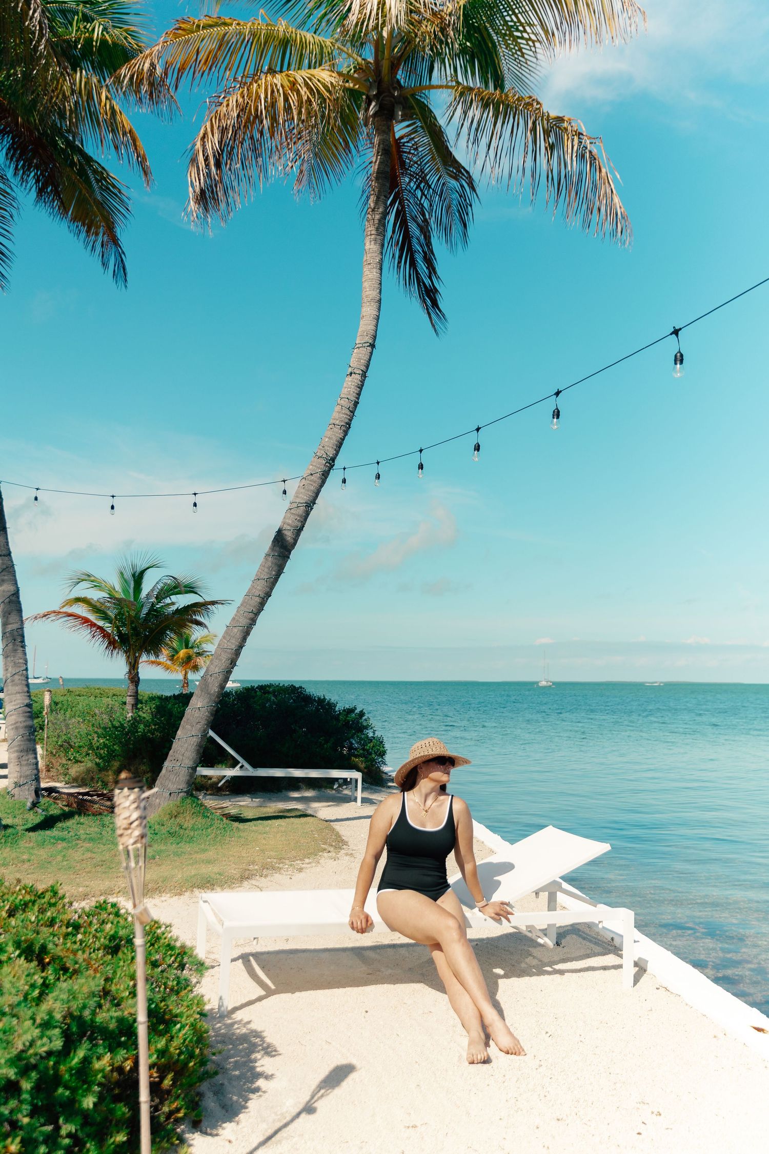 Woman in black swimsuit and hat relaxing on white lounge chair by tropical ocean with palm trees.