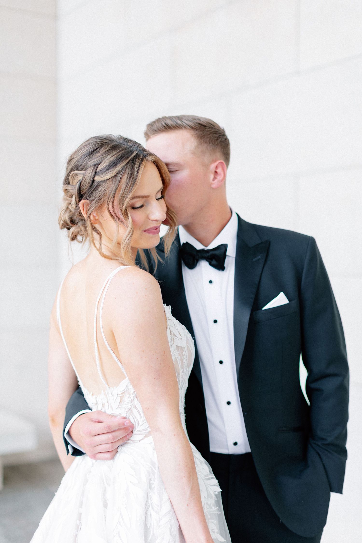Groom in black tuxedo kissing bride on forehead, bride wearing elegant backless white wedding gown.