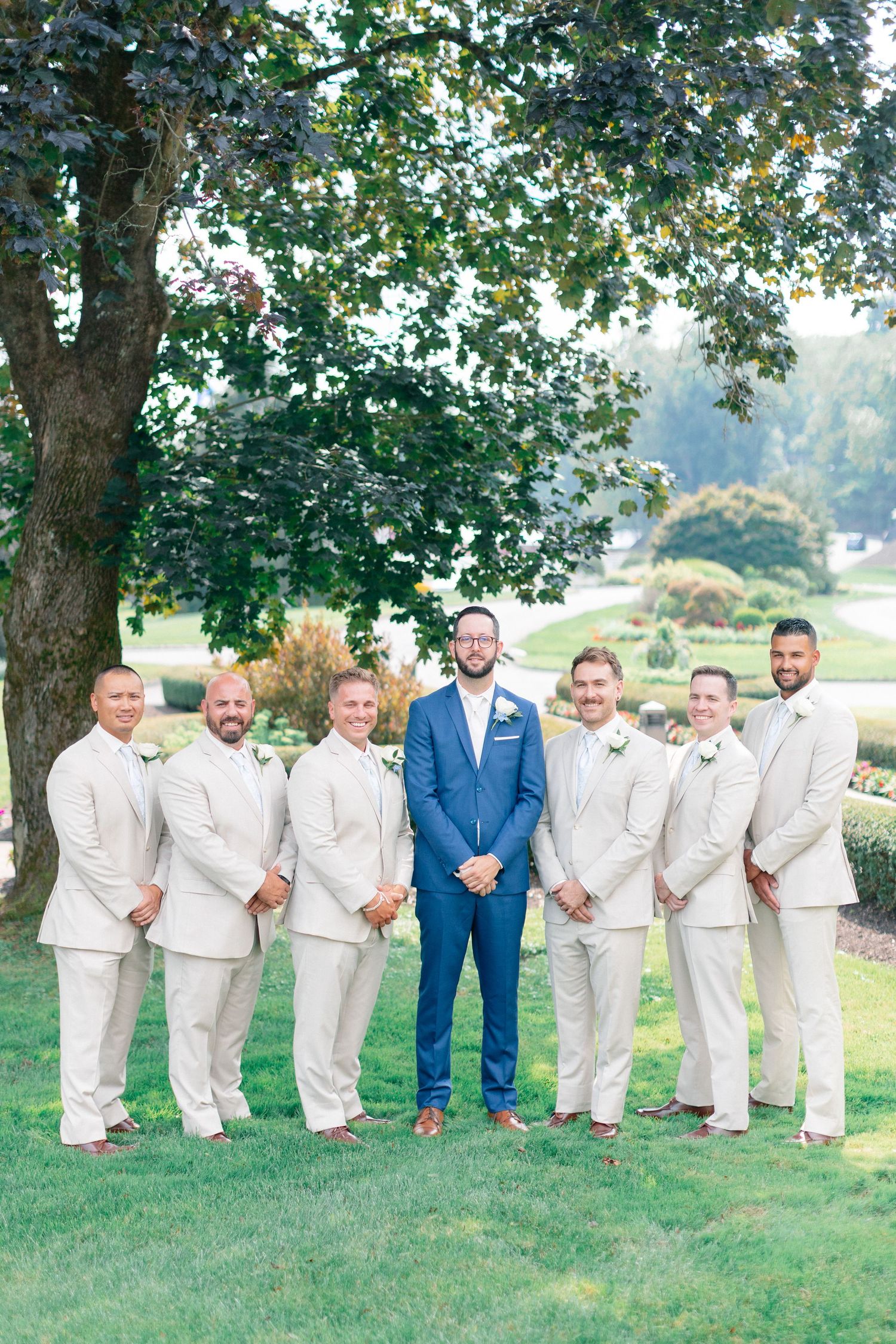 Groom in navy blue suit stands with six groomsmen in tan suits outdoors on a lush green lawn.