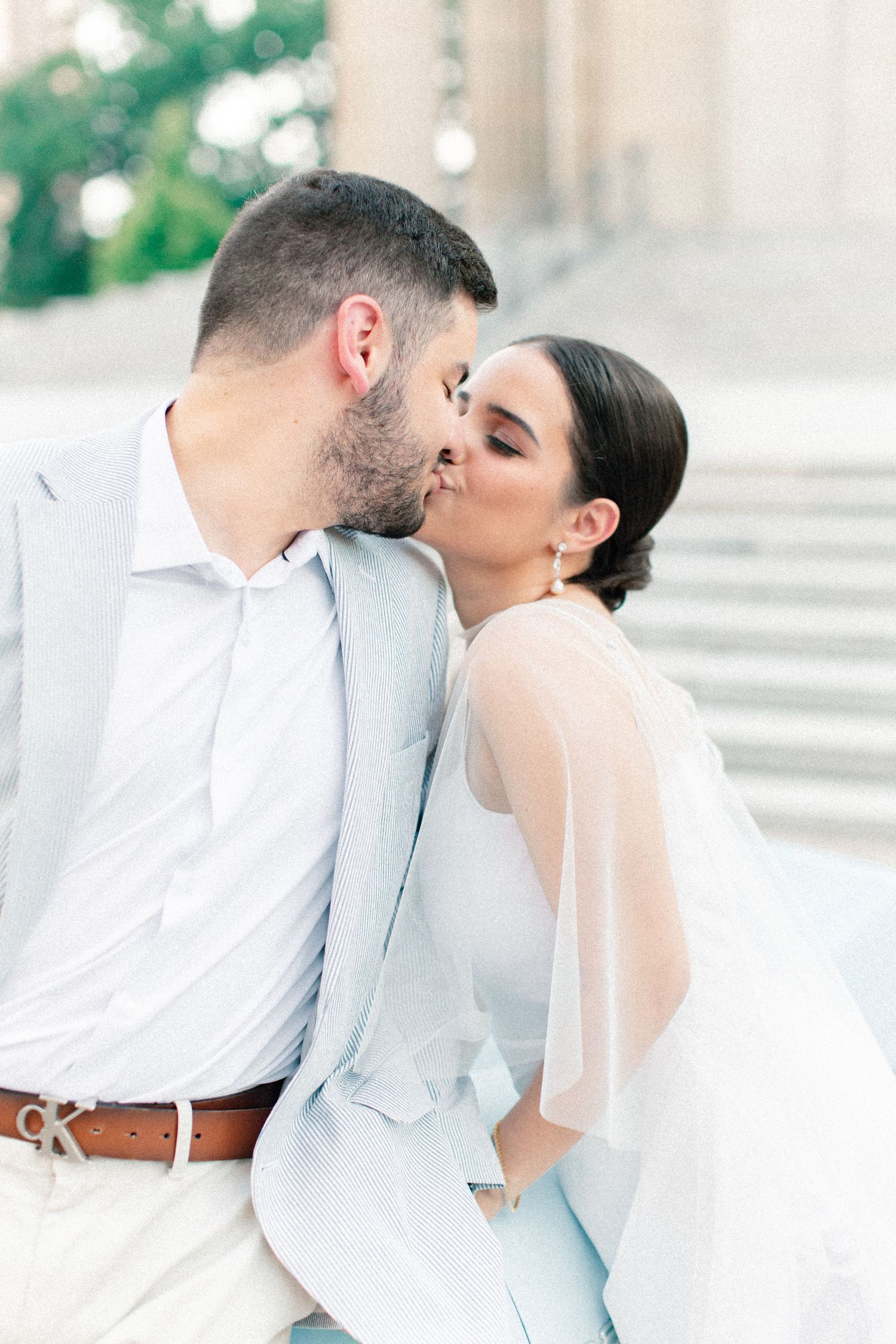 A couple shares a romantic kiss on outdoor steps, the groom in a light blue suit and bride in a white gown with sheer sleeves.