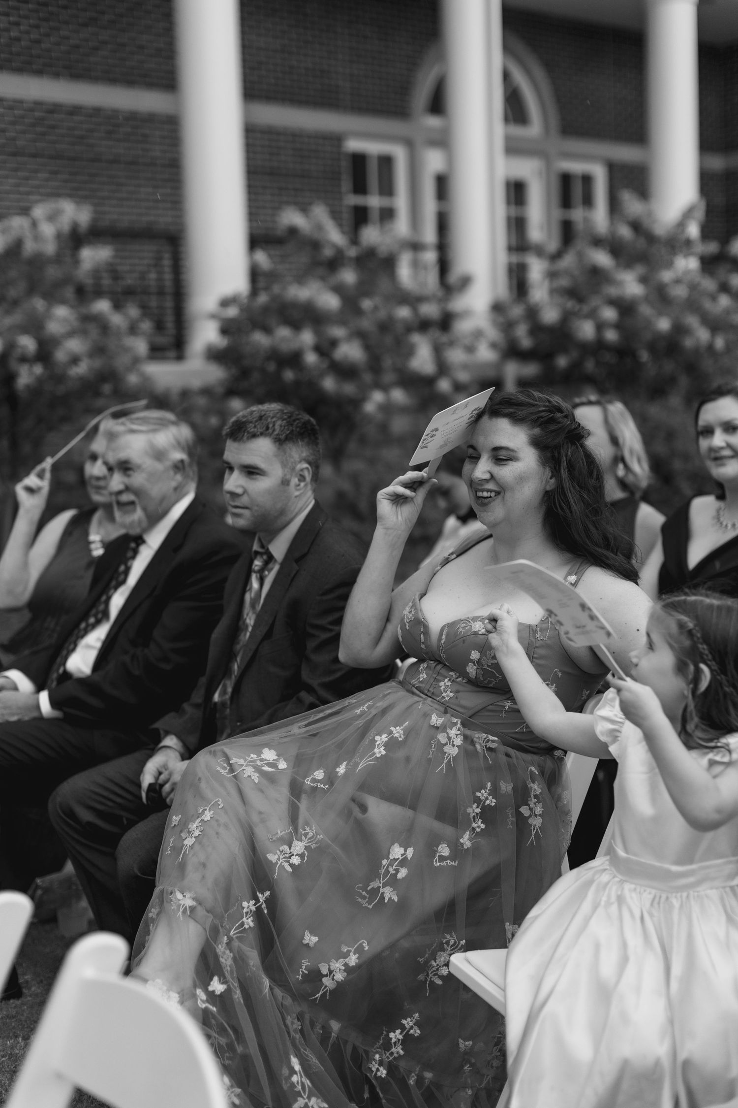 Black and white image of laughing wedding guests fanning themselves at outdoor summer ceremony.