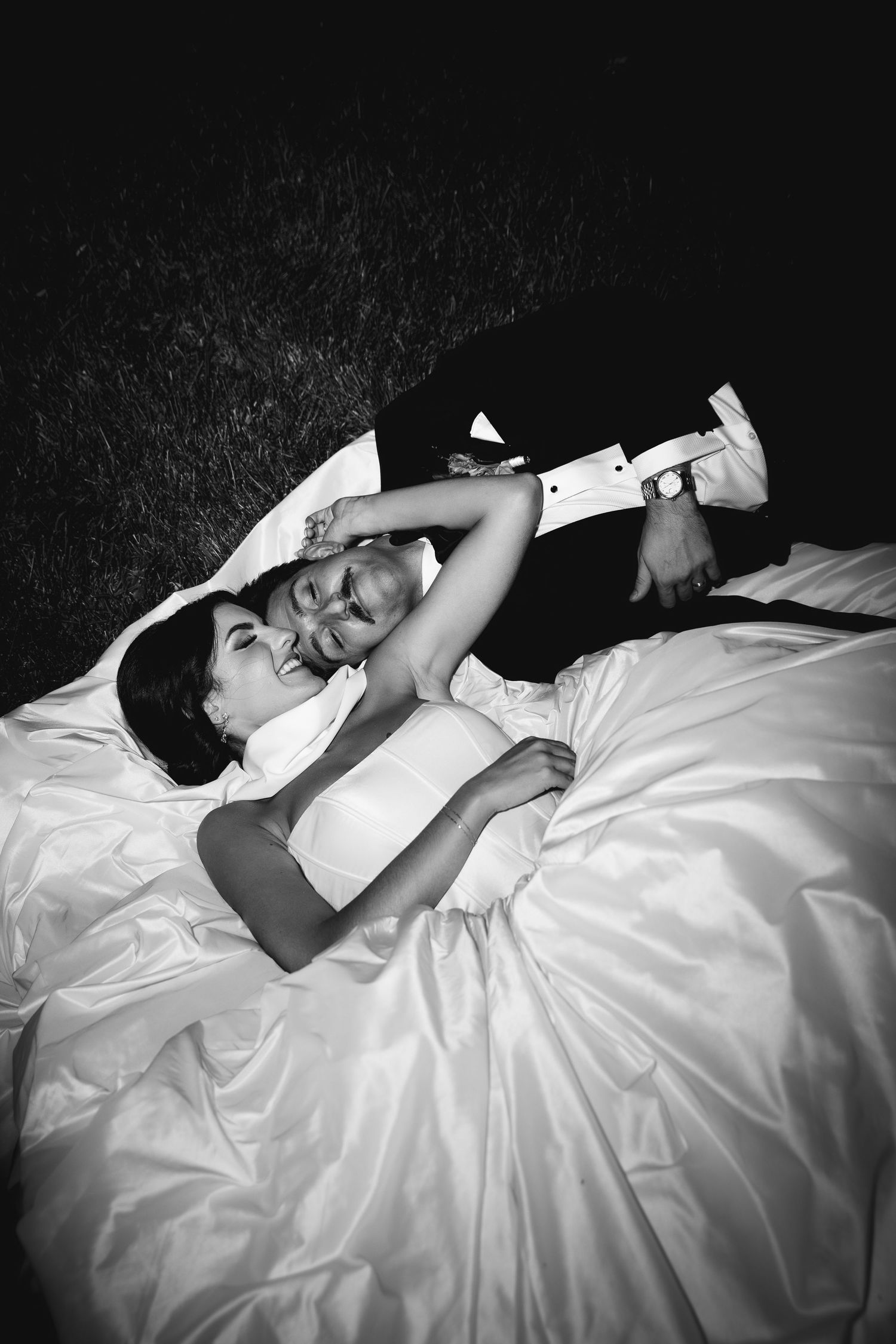 Black and white photo of a laughing bride and groom lying together on a white wedding dress at night.