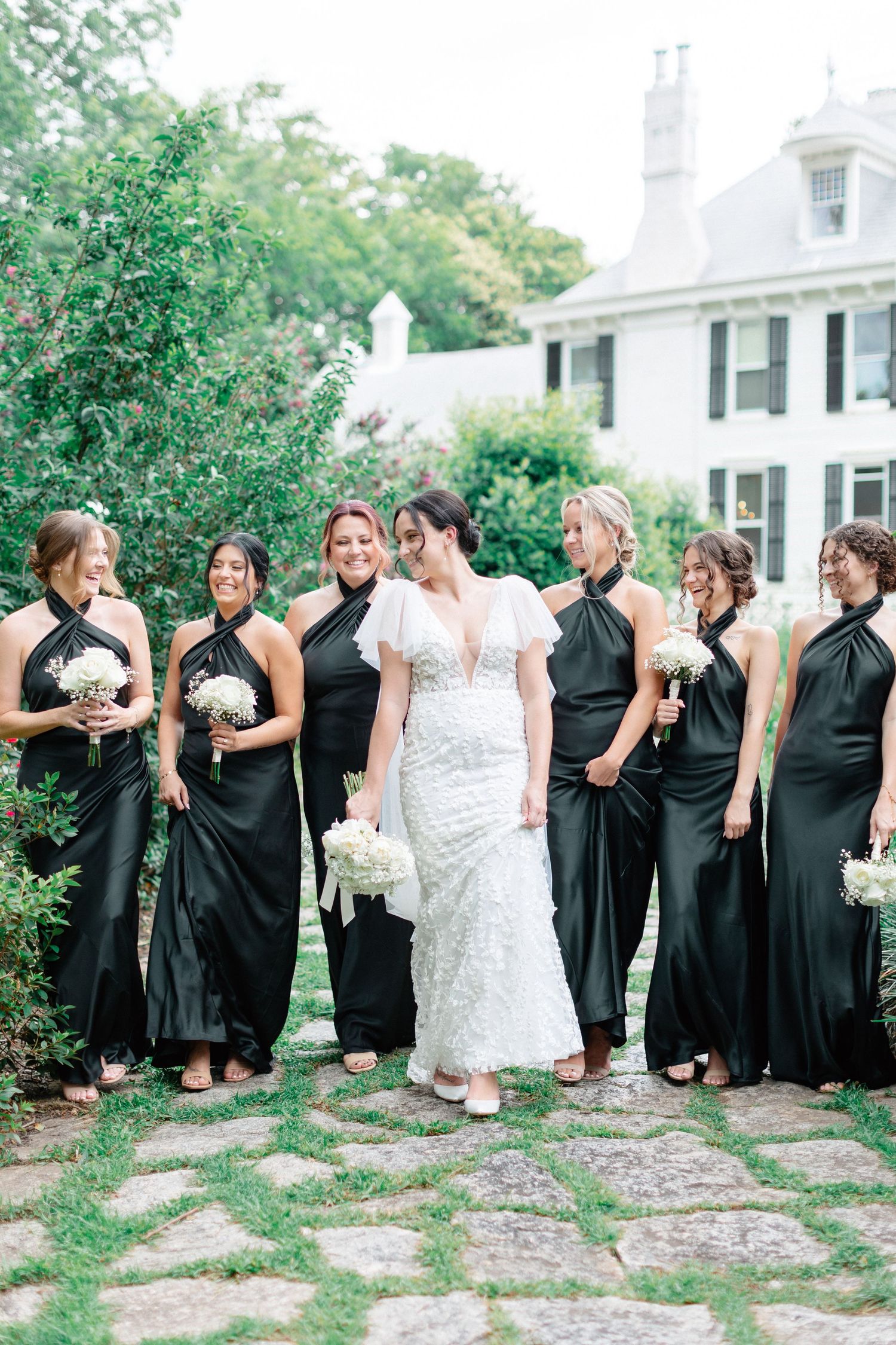 Bride in white lace gown smiles with six bridesmaids in black halter dresses on cobblestone path.