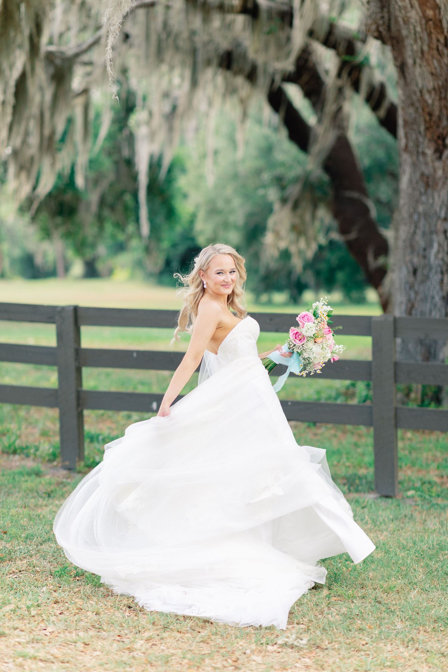 Smiling bride in flowing white gown twirling outdoors near rustic fence and moss-draped oak tree, holding colorful bouquet, Bella Cosa
