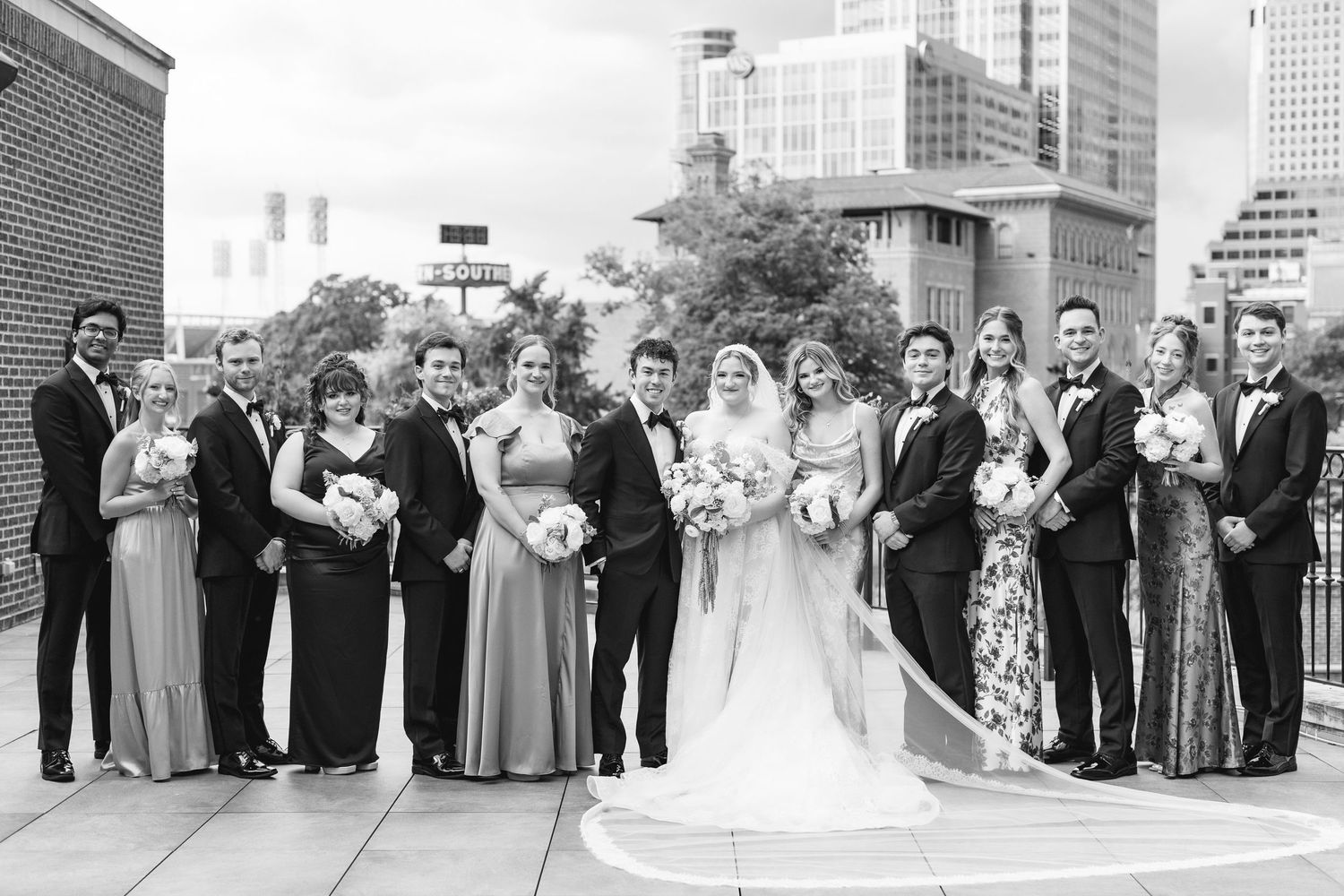 Black and white wedding party photo on rooftop with city skyline, bride in white gown surrounded by bridesmaids and groomsmen.