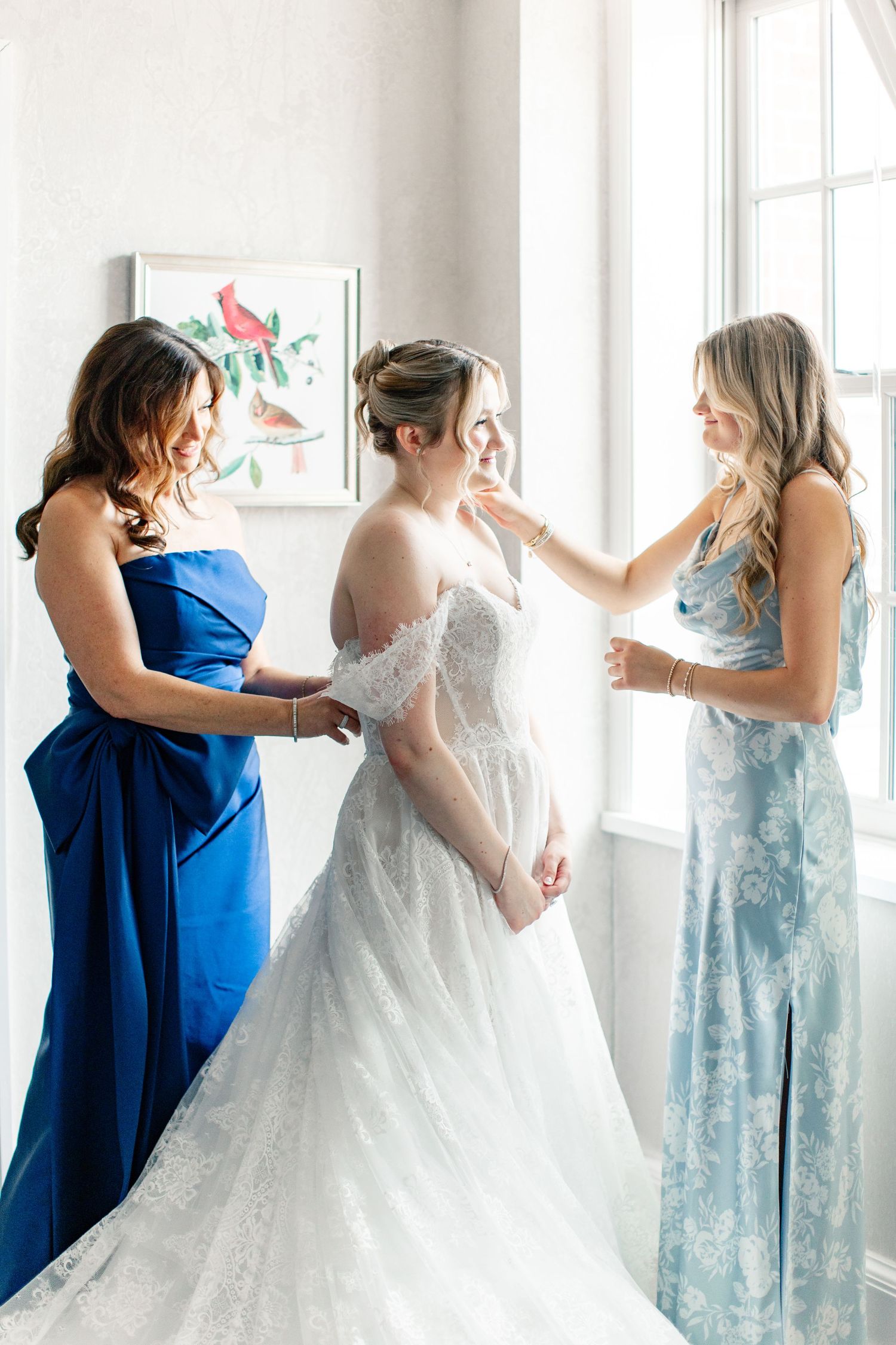 Two bridesmaids in blue dresses help a bride in a white lace gown get ready near a bright window.