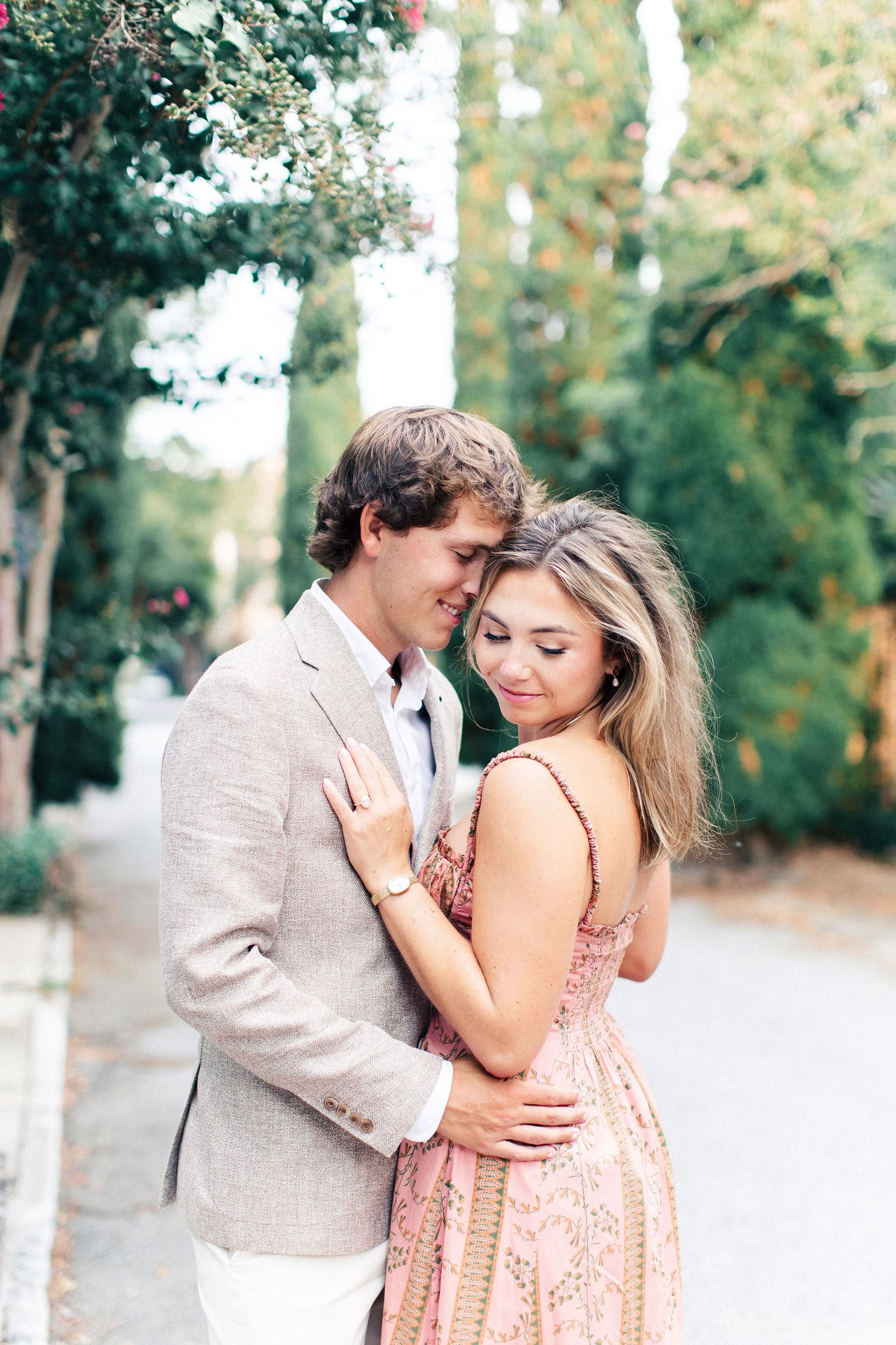 A couple embraces tenderly on a tree-lined street, the woman in a pink dress and man in a beige suit.