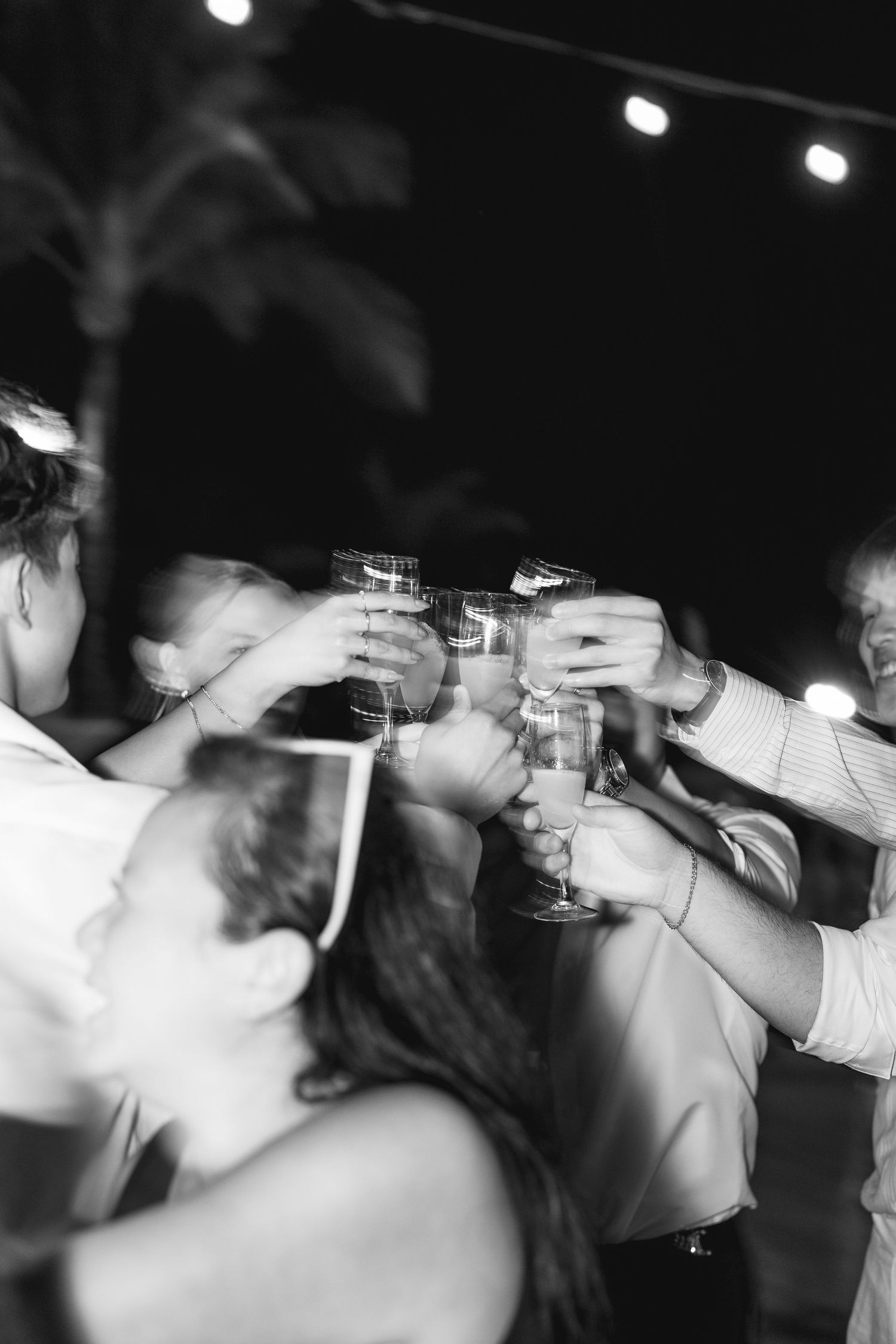 Black and white photo of wedding guests clinking champagne glasses in a celebratory toast under string lights.