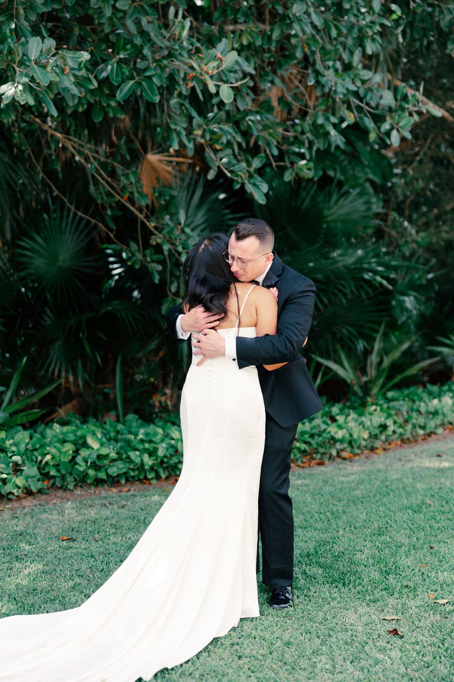A bride and groom share a tender embrace on a lush green lawn surrounded by tropical foliage on their wedding day.