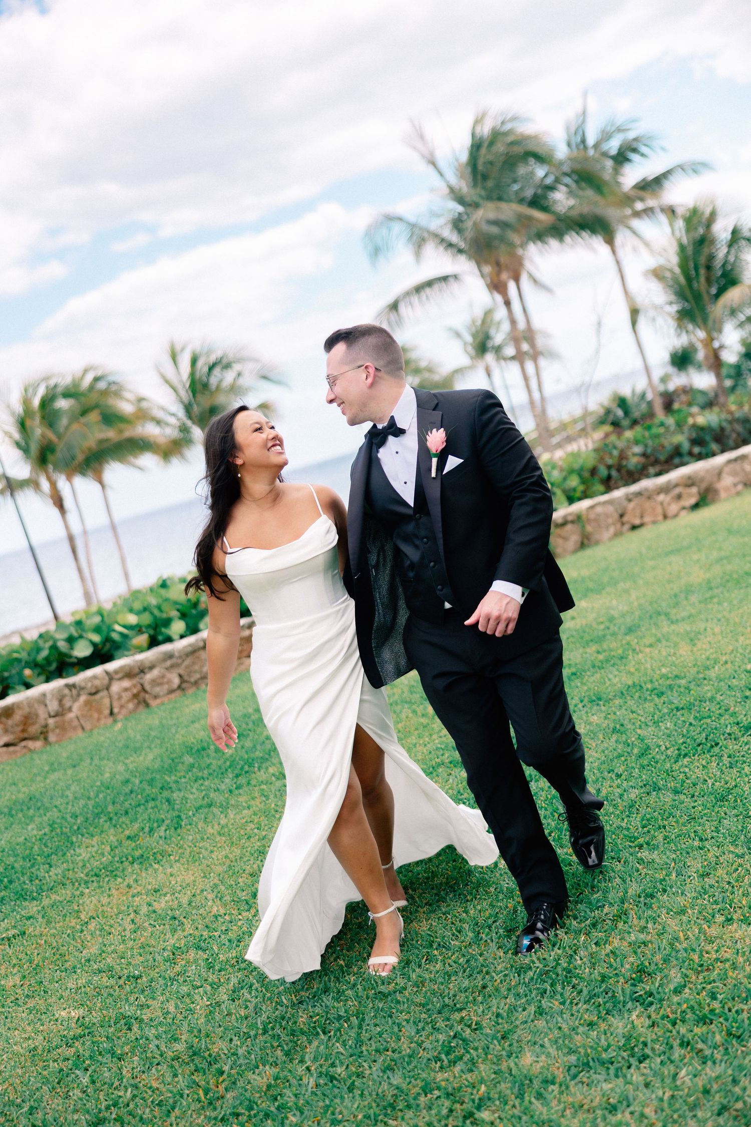 A joyful bride and groom walk together on lush green grass with palm trees and ocean in the background at a tropical wedding.