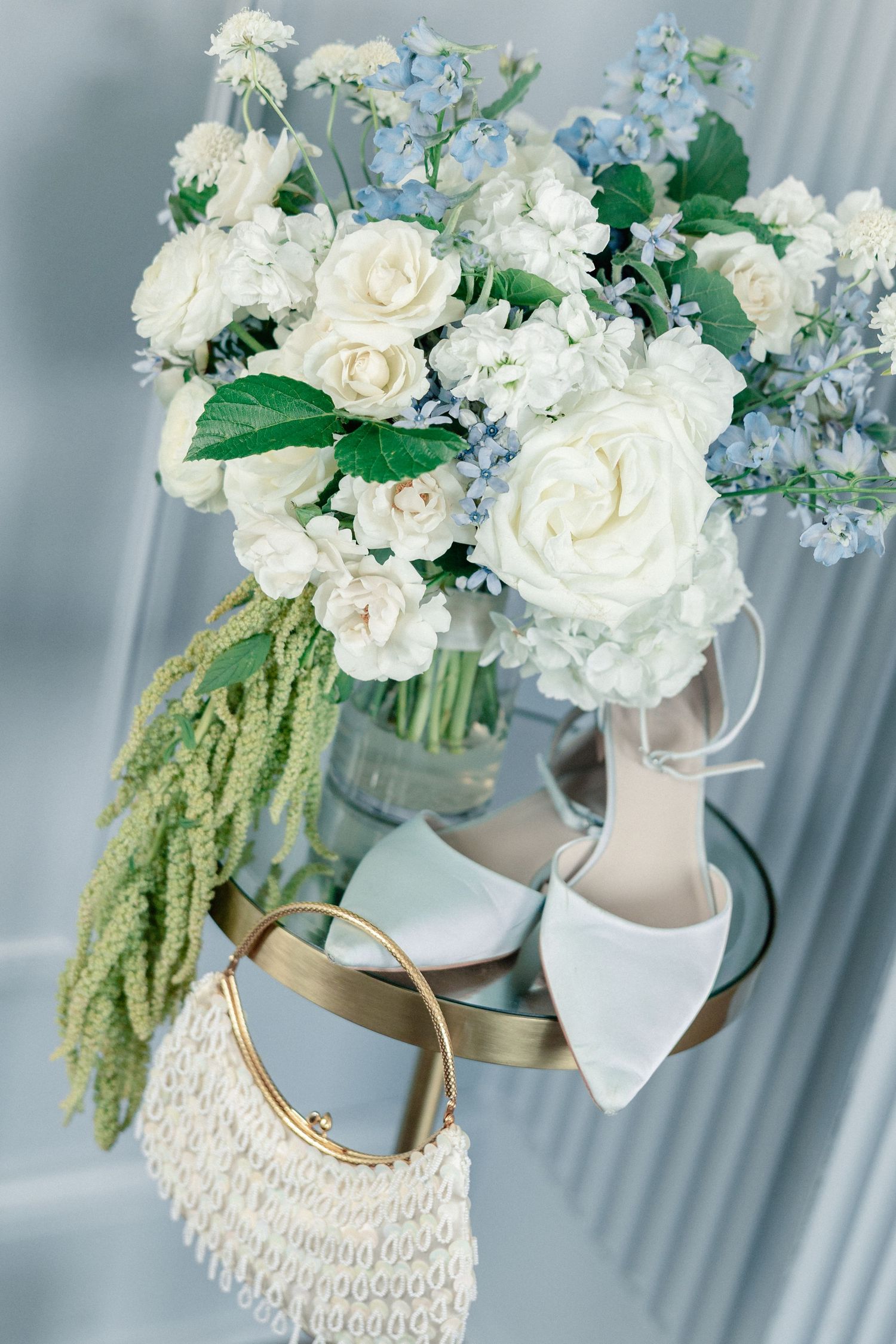 Elegant bridal flat lay with white roses, blue flowers, satin heels, and a pearl beaded clutch on a gold tray.