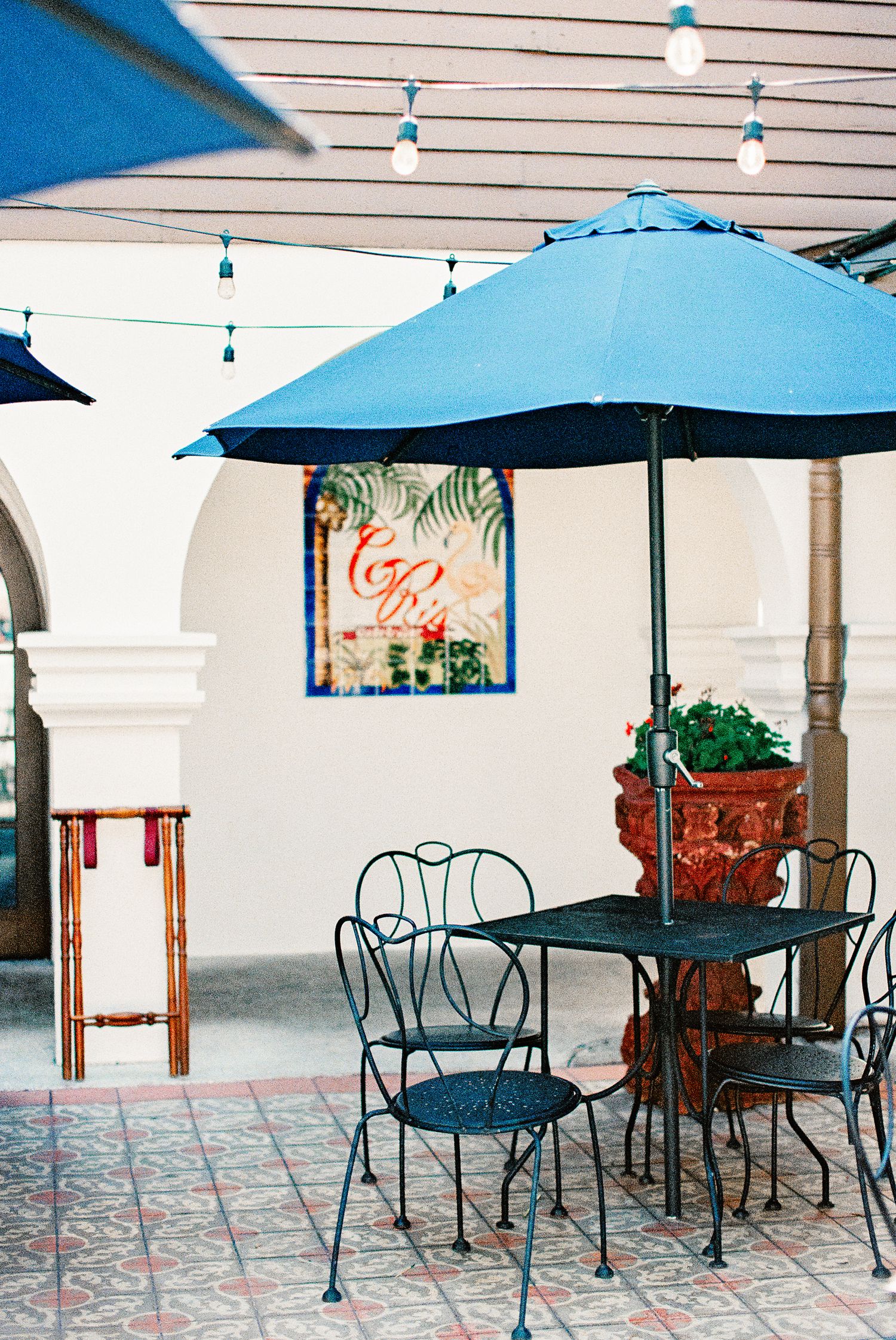 Blue patio umbrella shading wrought iron table and chairs on tiled outdoor terrace with string lights and tropical decor.