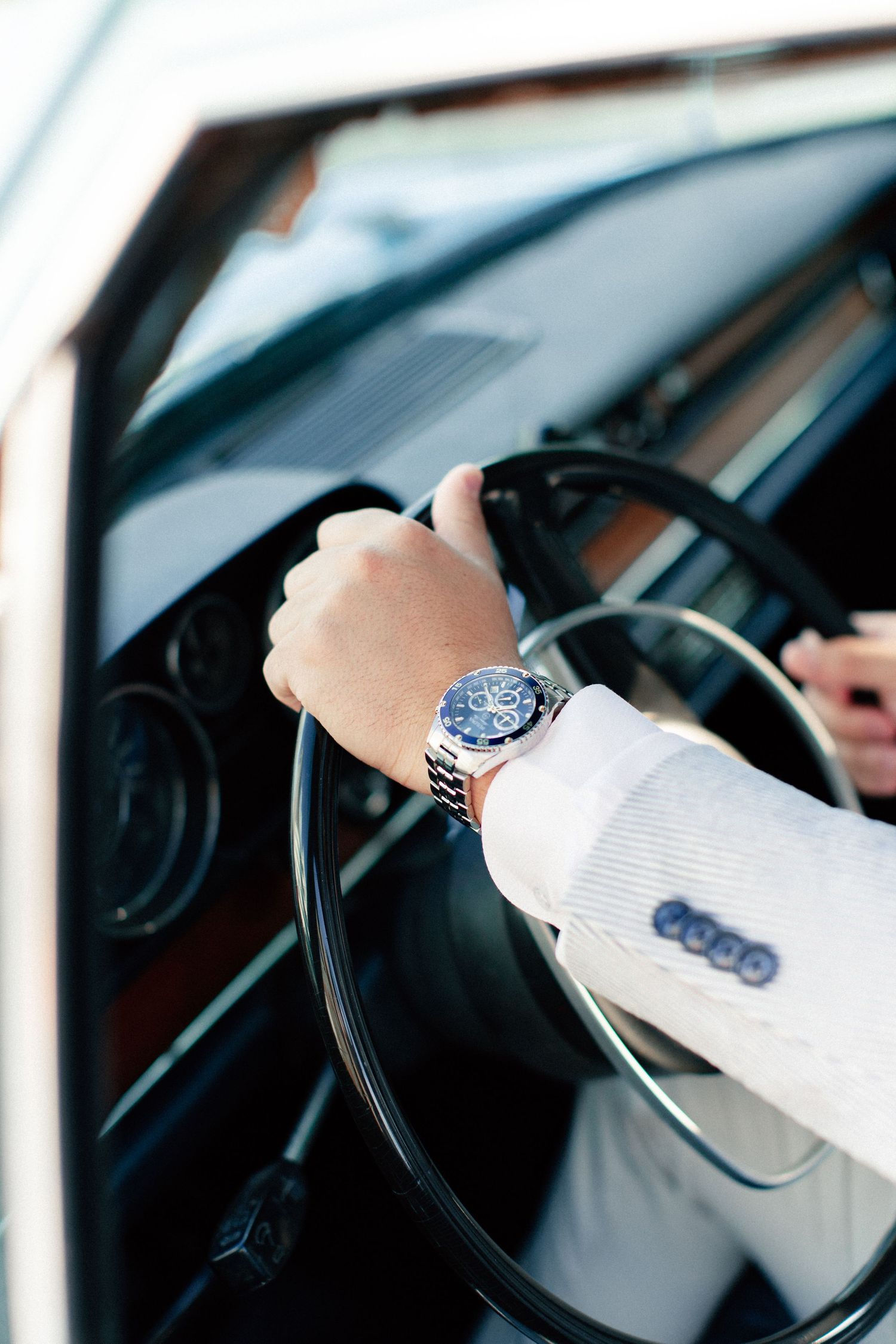 Close-up of a man in a white suit driving a classic car, wearing a luxury chronograph watch on his wrist.